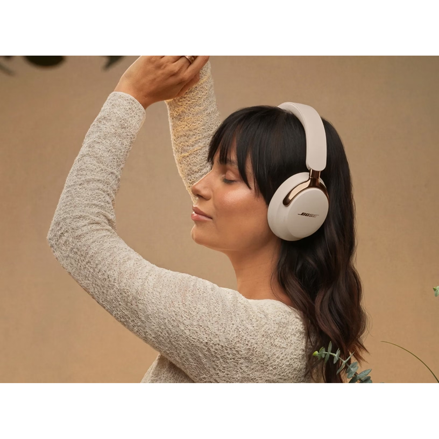 Woman with closed eyes wearing white headphones, raising arm, in front of a brown background. Bose logo on headphones.