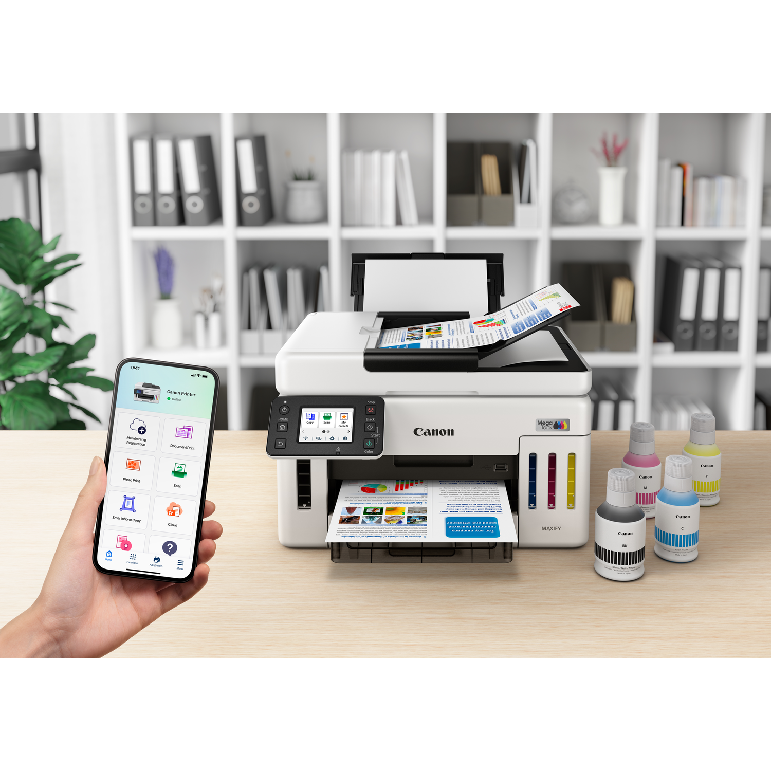 A Canon printer with a smartphone showing its app. Ink bottles are on the table. A hand holds the phone. Bookshelves are in the background.