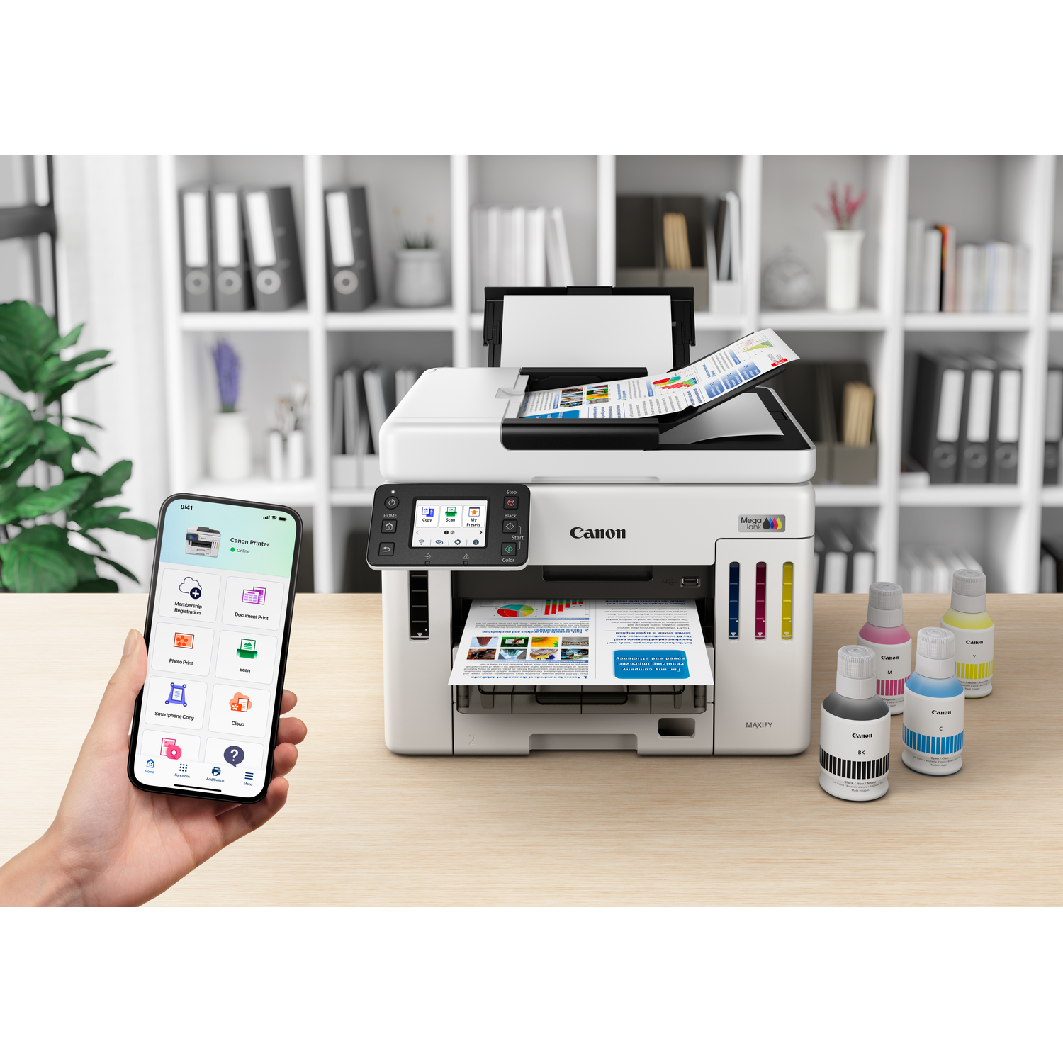 A Canon printer with a smartphone showing an app. Ink bottles are on the right. Background shows a shelf with books.