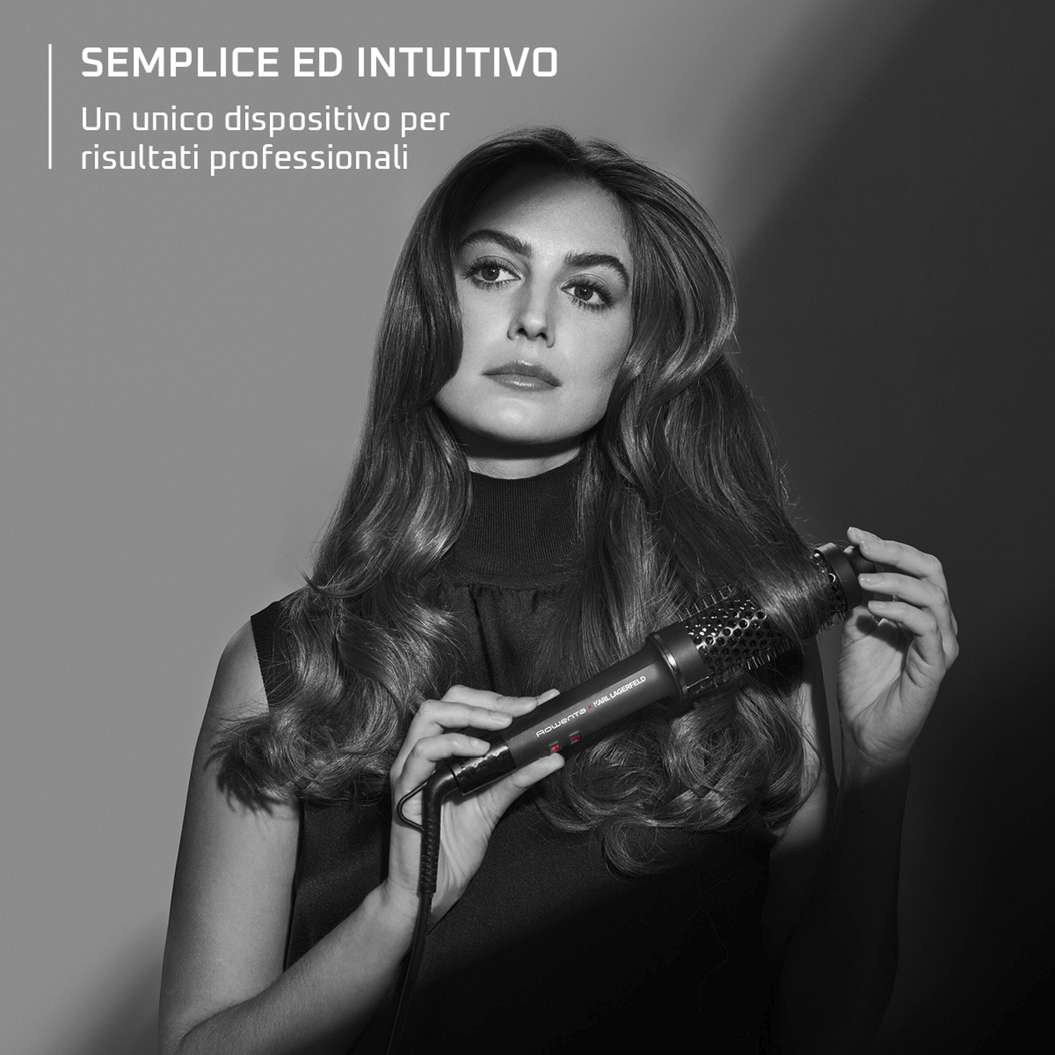 Una donna con i capelli lunghi usa una spazzola per lo styling. Tiene la spazzola e lo sfondo è un gradiente di grigio.