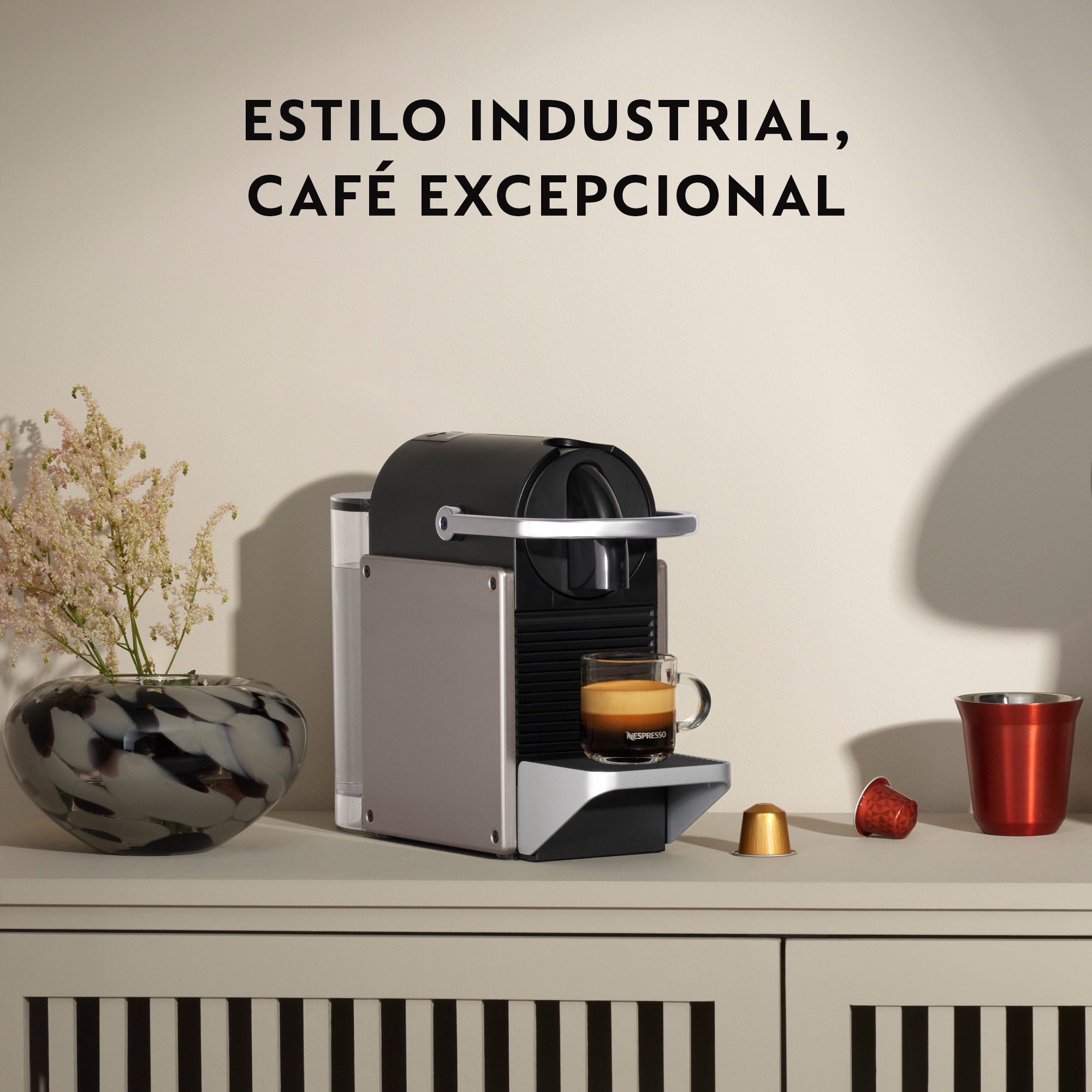 Una máquina Nespresso con una taza de café, cápsulas y una taza roja sobre una superficie blanca. Texto: Estilo industrial, café excepcional.
