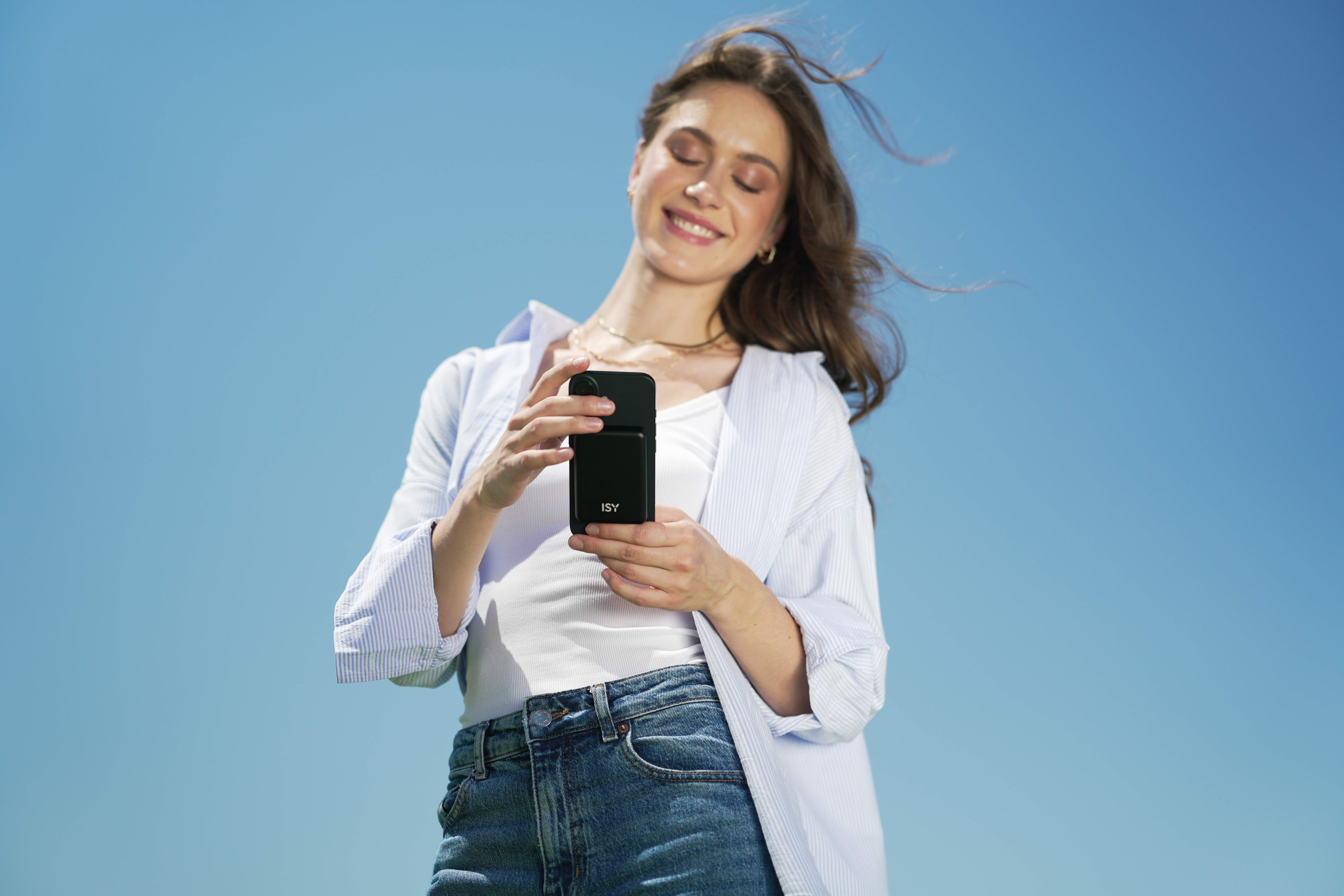 Donna che tiene in mano un dispositivo nero contro un cielo blu. Indossa una camicia bianca e jeans, sorridendo.
