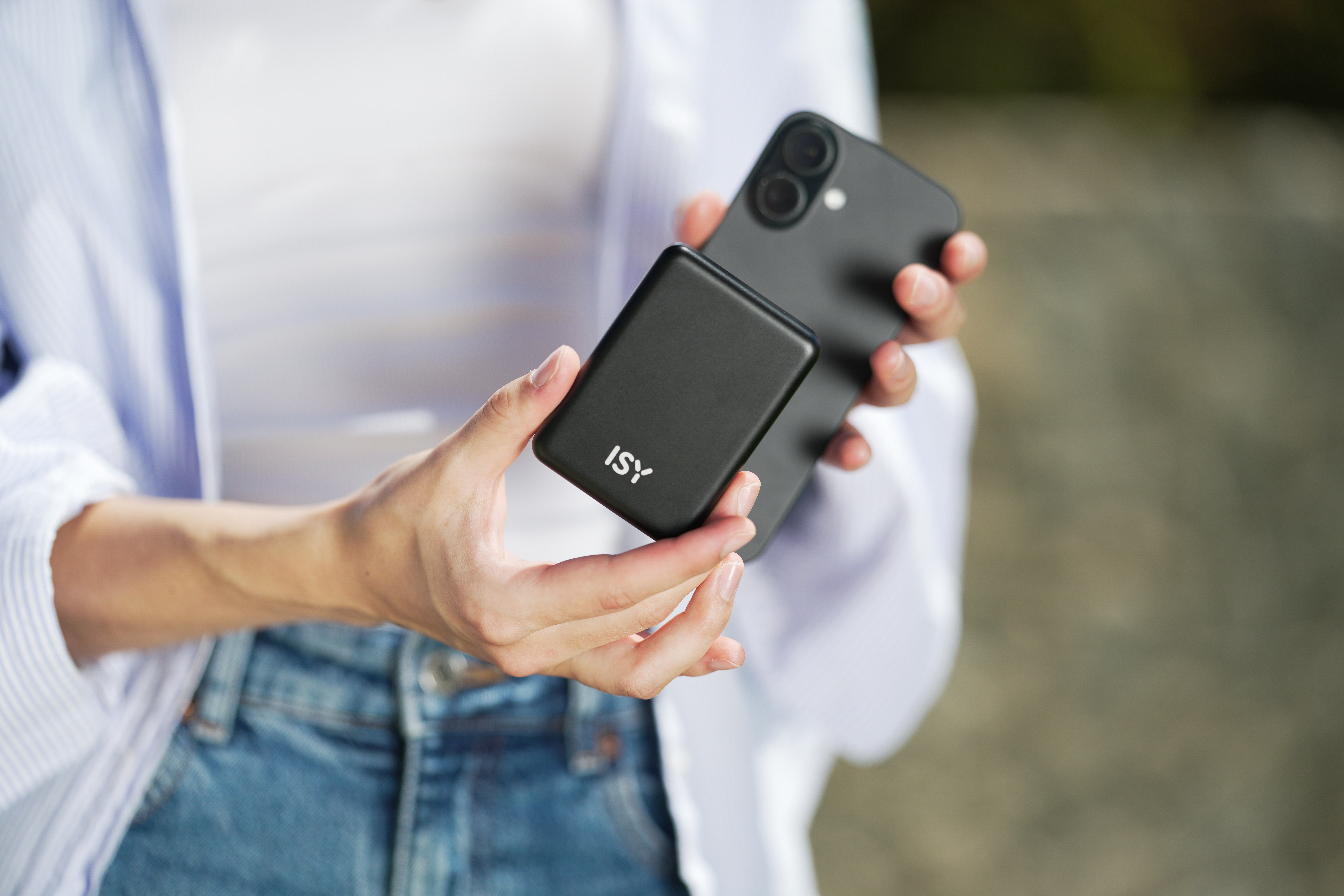 Una persona tiene in mano un telefono nero e un power bank nero con "ISY", indossa una camicia bianca e jeans.