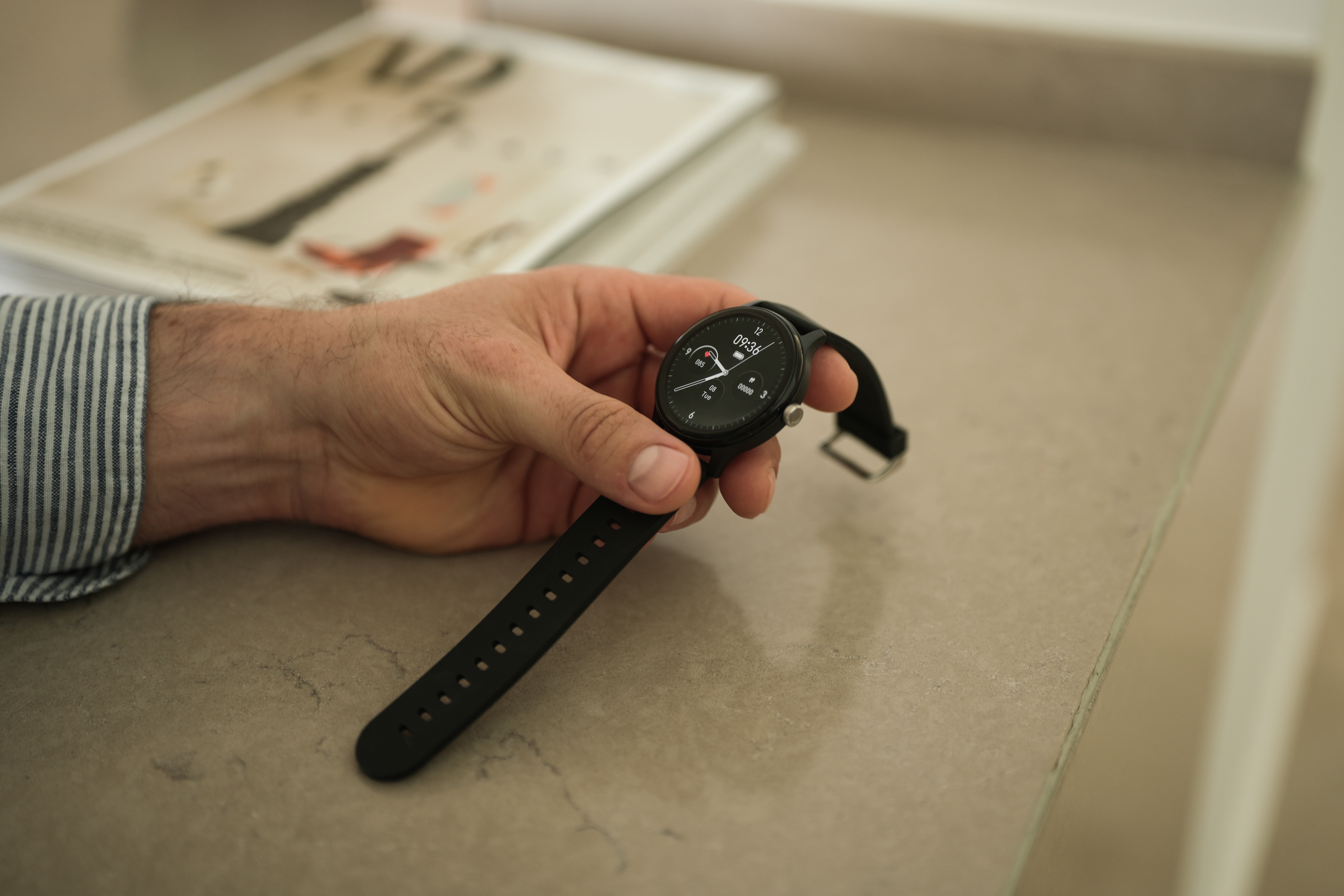 Die Hand einer Person hält eine schwarze Smartwatch. Die Uhr zeigt die Zeit an. Im unscharfen Hintergrund befindet sich eine Zeitschrift.