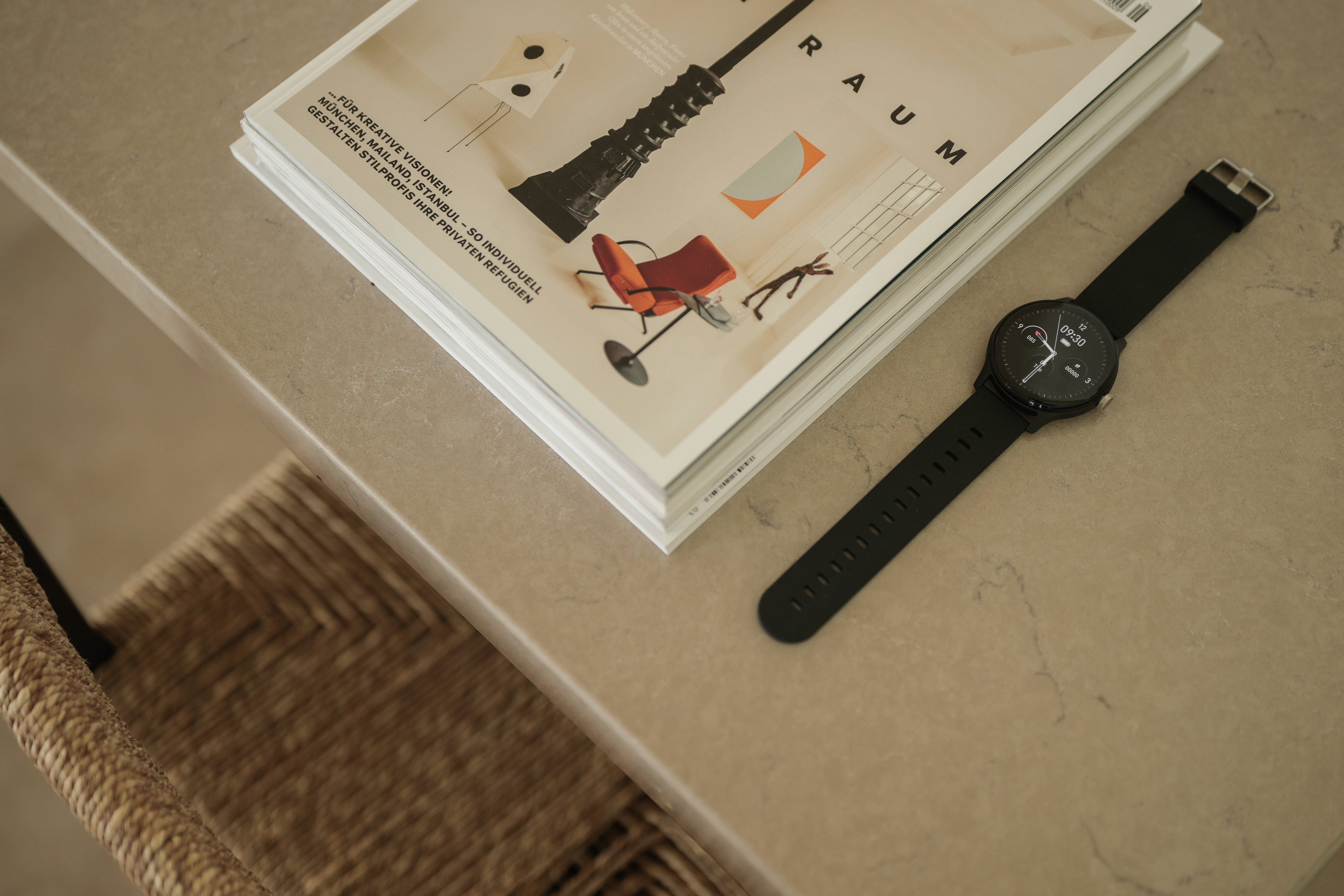 Eine Uhr liegt neben einer Zeitschrift auf einem Tisch. Ein Stuhl ist teilweise sichtbar. Die Uhr zeigt 09:30.