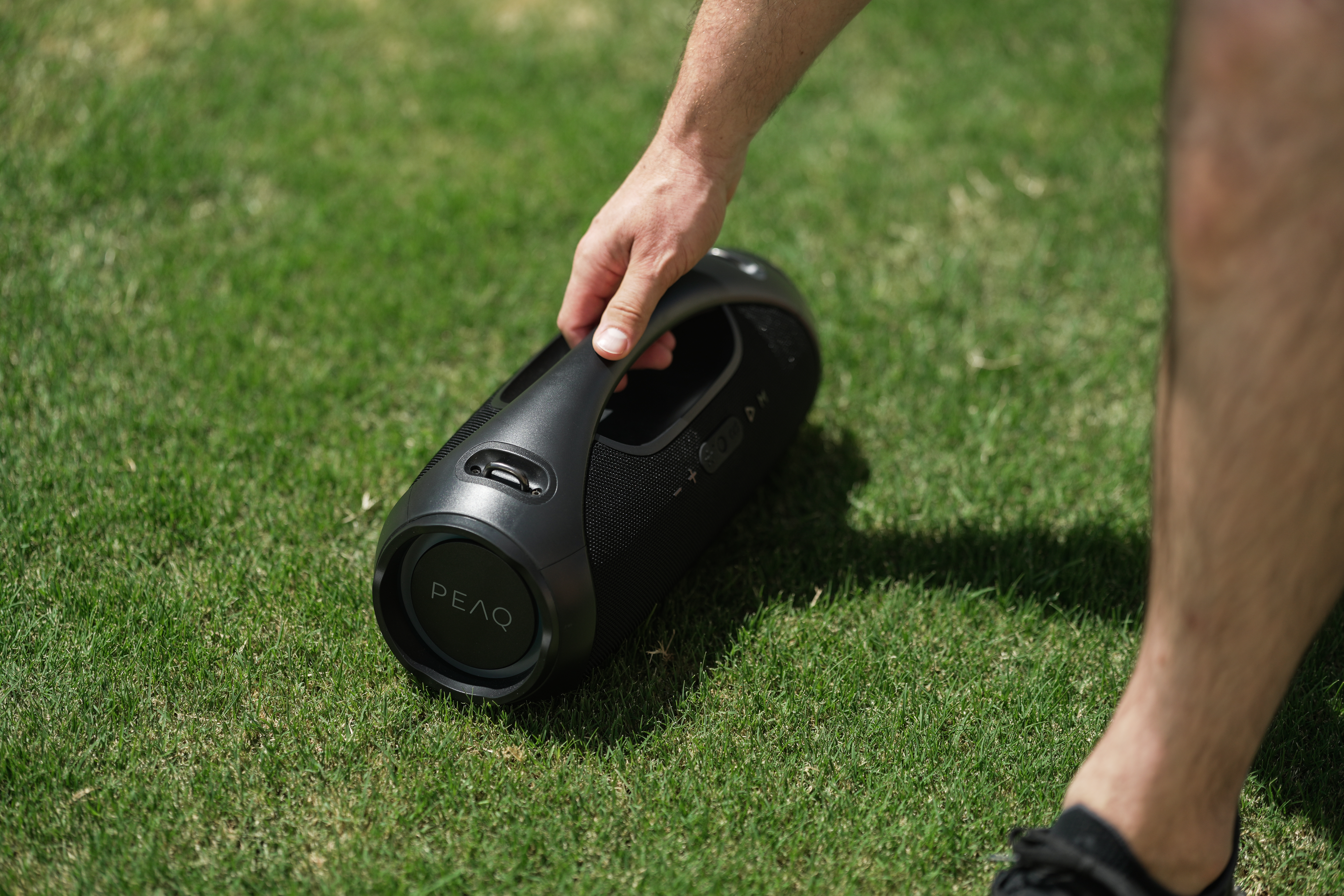 La main d'une personne tient un haut-parleur noir sur de l'herbe verte. Le haut-parleur porte le mot PEAQ.