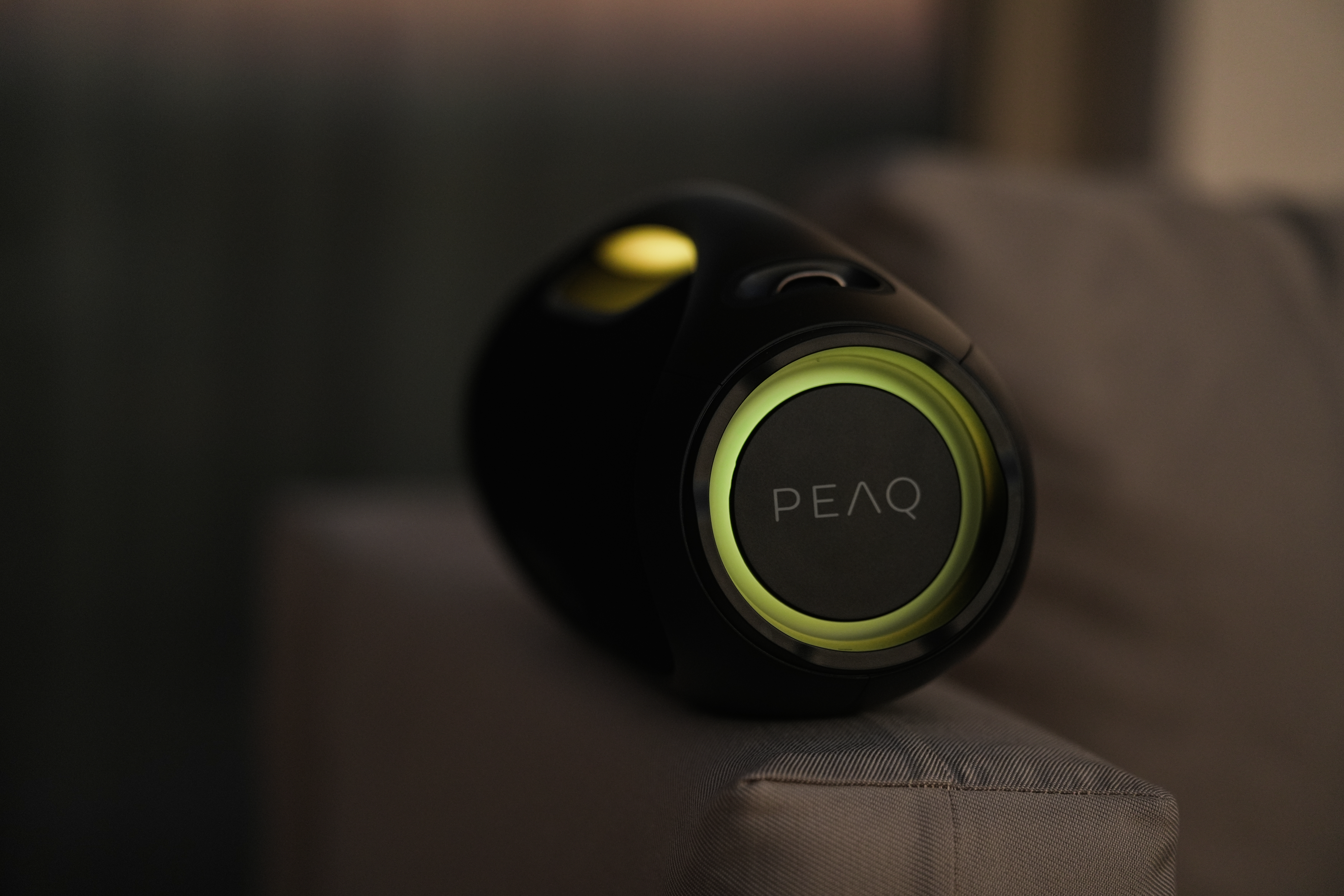 Un appareil noir avec 'PEAQ' dessus, avec des lumières vertes et jaunes. Il est sur un coussin gris.