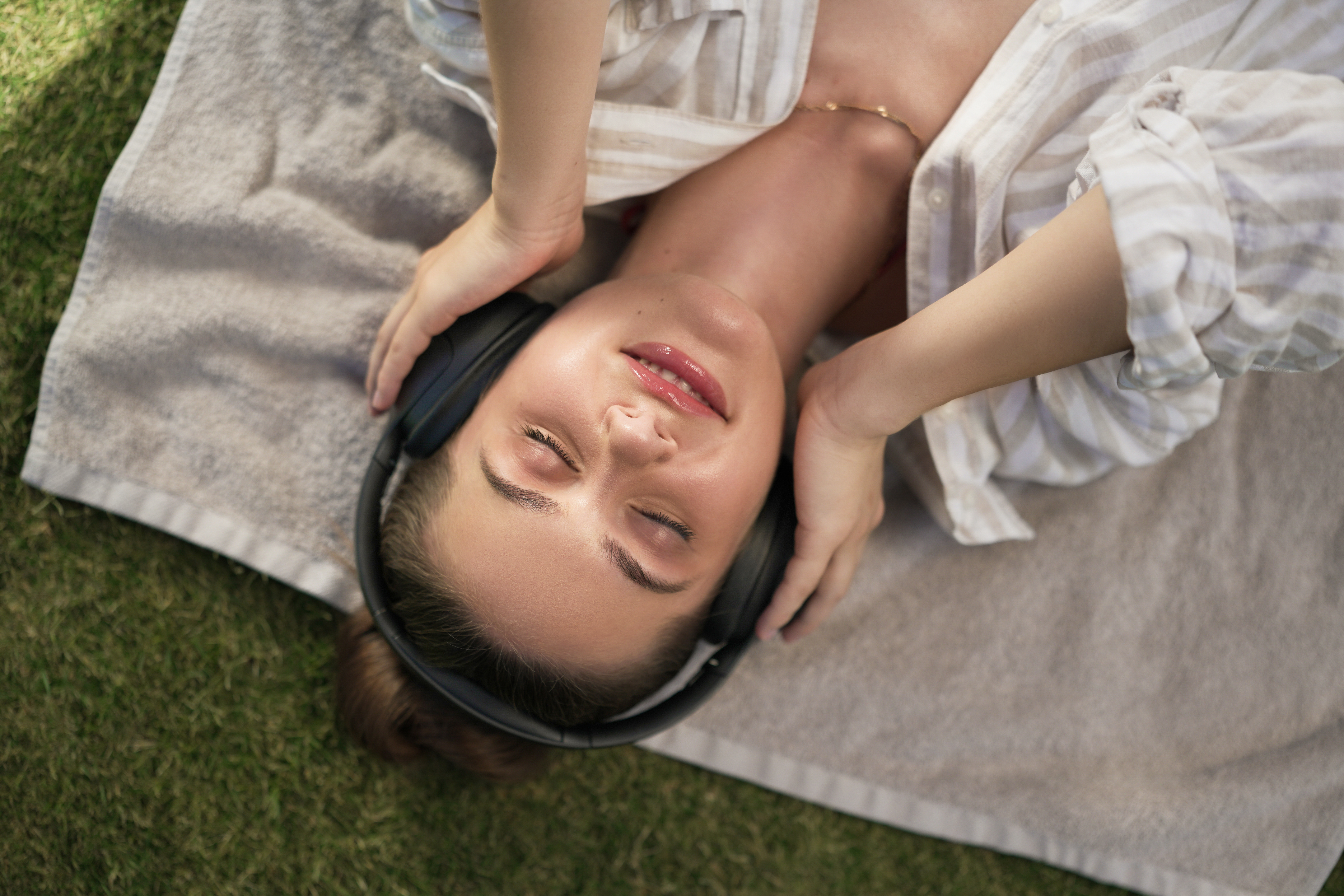 Une femme avec un casque est allongée sur une serviette sur l'herbe. Elle porte une chemise rayée et a les yeux fermés.