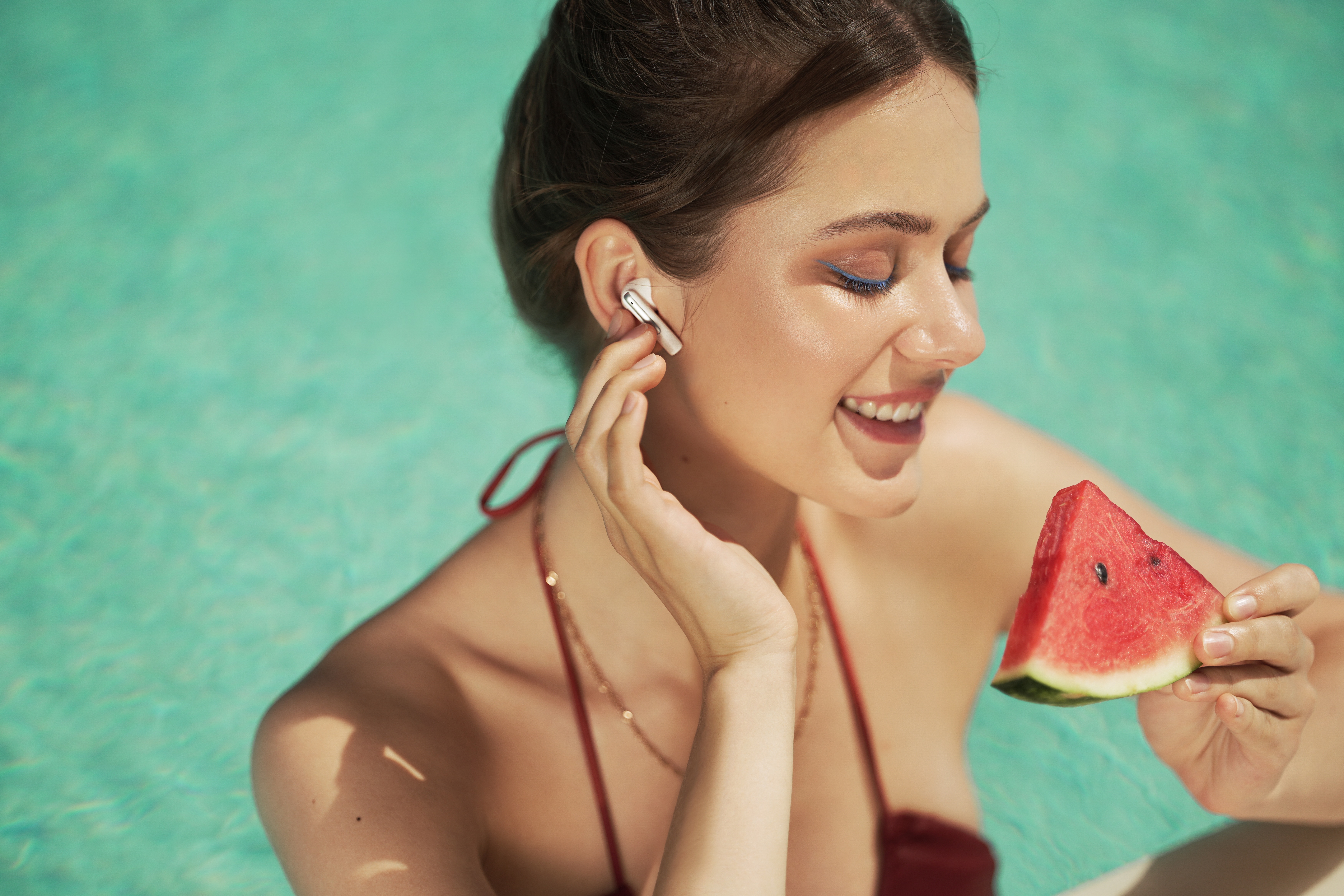 Femme dans une piscine tient une tranche de pastèque, avec des écouteurs. Eyeliner bleu, haut de bikini rouge et collier doré.
