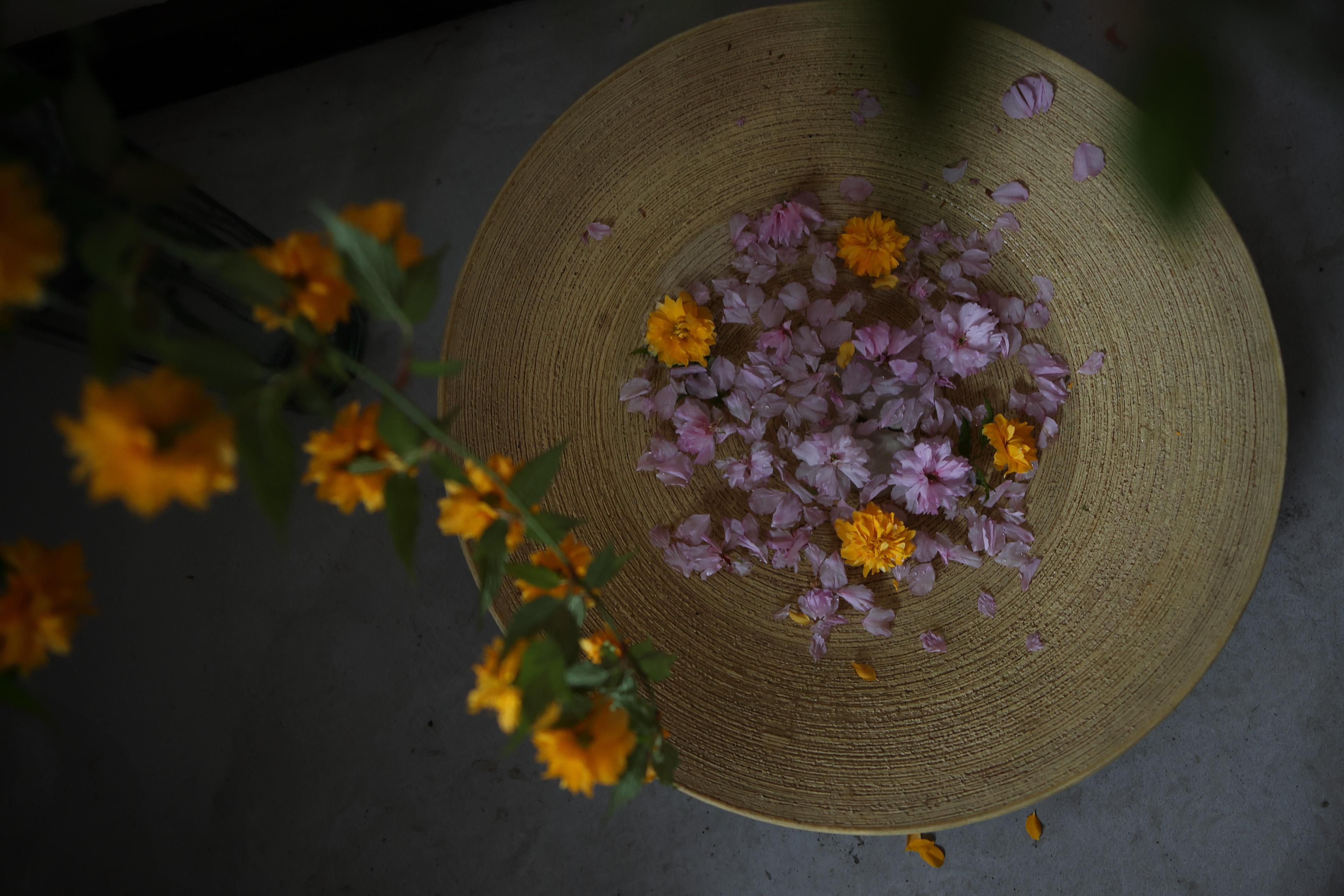 Eine runde, goldene Schale mit rosa und gelben Blüten und Blütenblättern. Gelbe Blumen an der Seite.