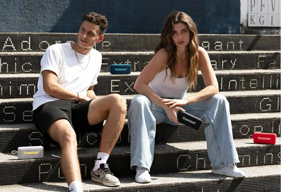 Dwie osoby siedzą na schodach z głośnikami. Mężczyzna w białym t-shircie i czarnych spodenkach, kobieta w białym topie i dżinsach.