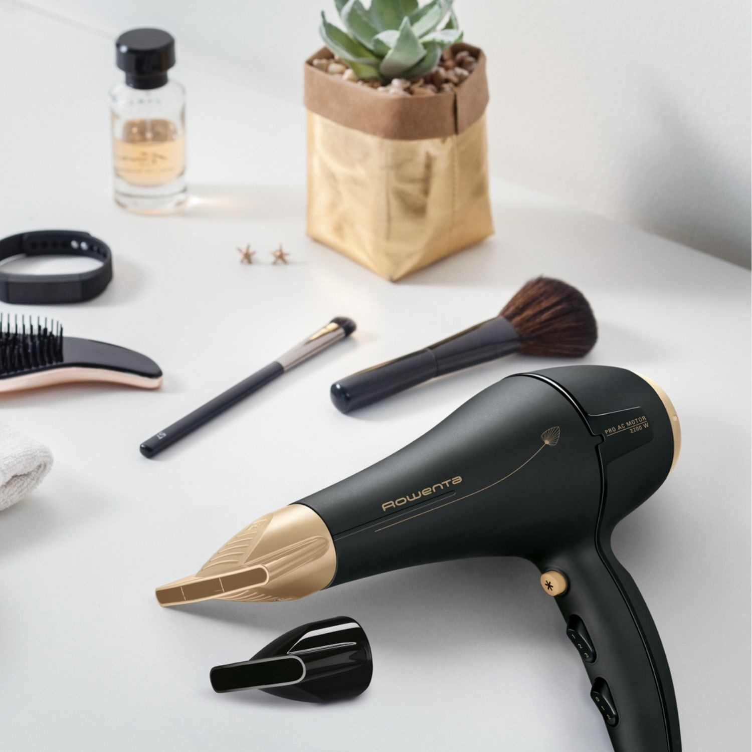 Un asciugacapelli nero e oro con pennelli e accessori per il trucco su una superficie bianca. C'è anche una pianta e profumo.