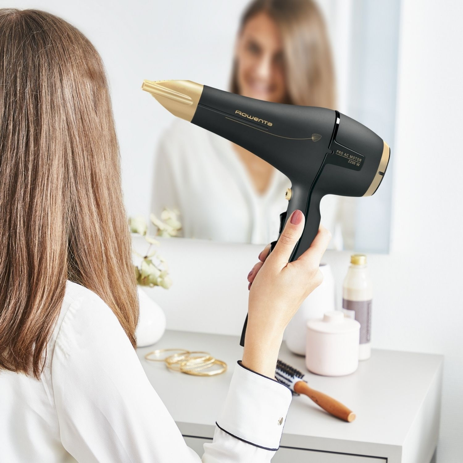 Una donna tiene un asciugacapelli nero e oro davanti a uno specchio. I capelli vengono acconciati. Sullo sfondo, un tavolo con accessori.