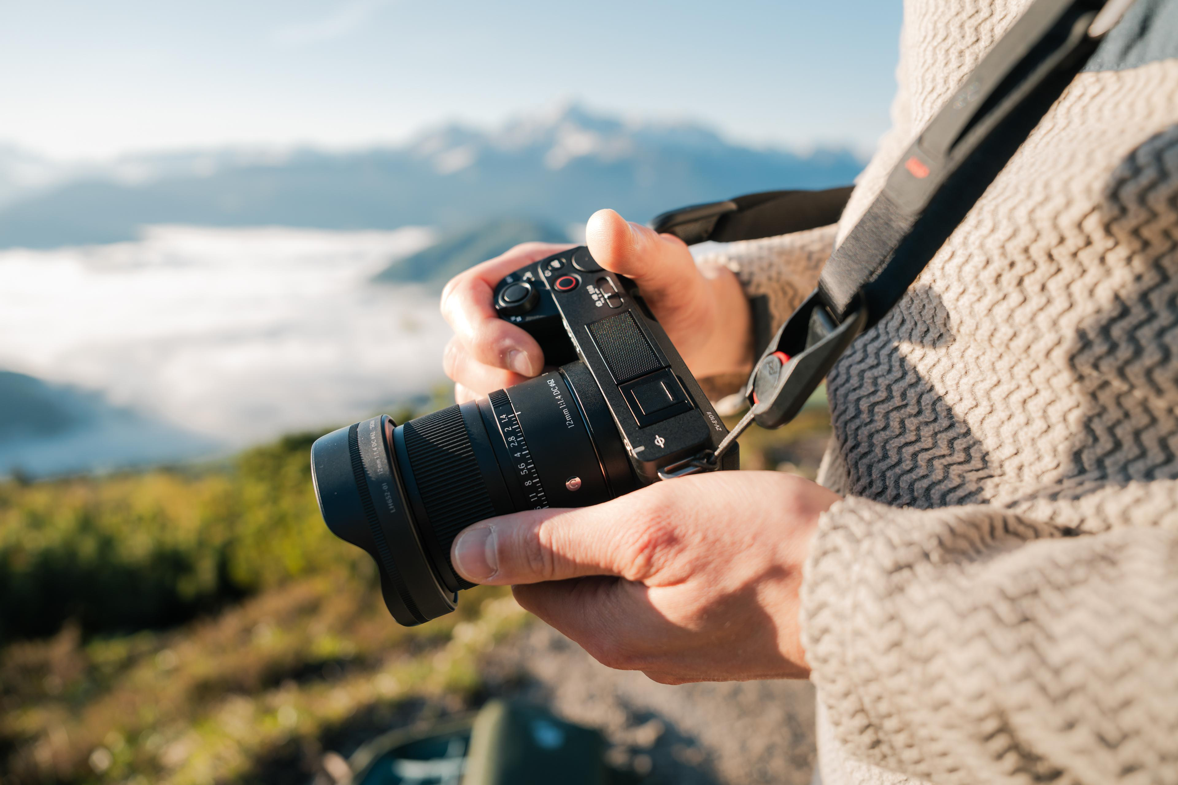 Eine Person, die eine schwarze Kamera mit einem Objektiv hält, mit einem malerischen Bergblick im Hintergrund.