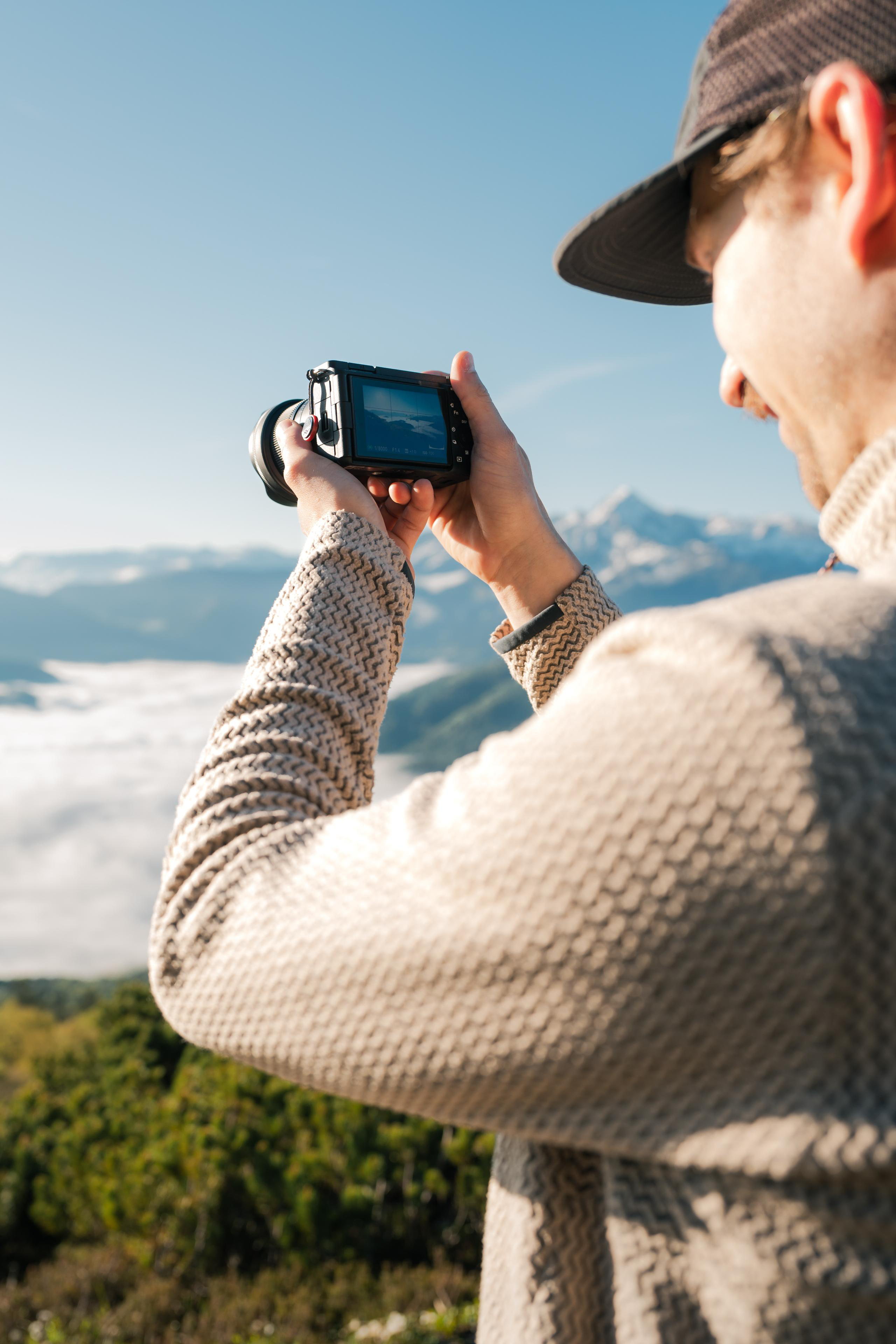 Eine Person, die eine Kamera hält und ein Bergpanorama fotografiert. Die Person trägt einen Hut und einen Pullover.