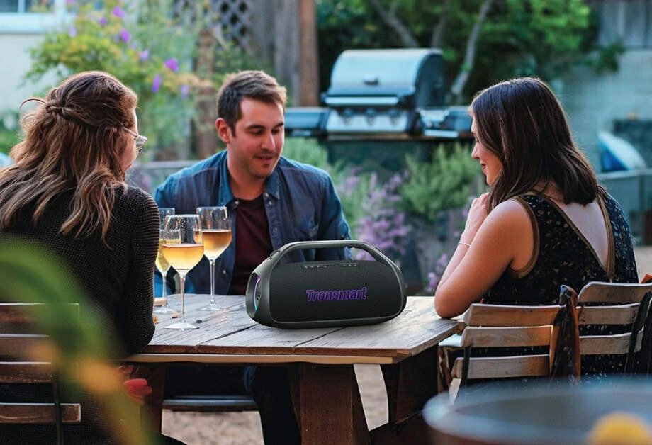 Trzy osoby siedzą przy stole z kieliszkami wina i głośnikiem. Grill w tle.