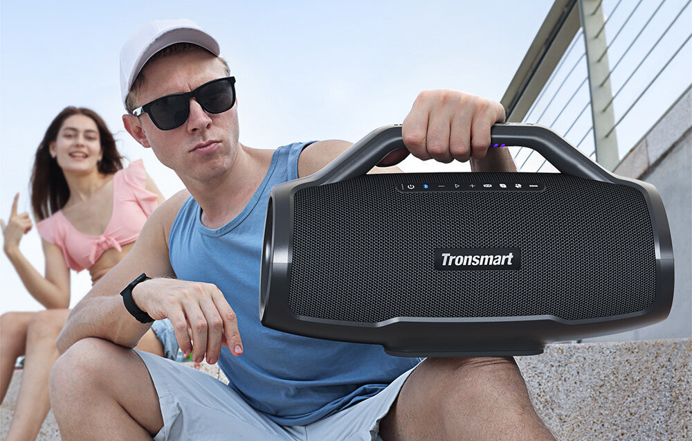 Mężczyzna w okularach przeciwsłonecznych trzyma czarny głośnik. Kobieta gestykuluje w tle. Oboje są na zewnątrz, na schodach.