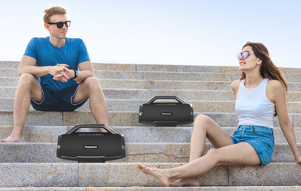 Dwie osoby siedzą na schodach z dwoma głośnikami. Mężczyzna w okularach przeciwsłonecznych, niebieskiej koszuli. Kobieta w okularach, białym topie i szortach.