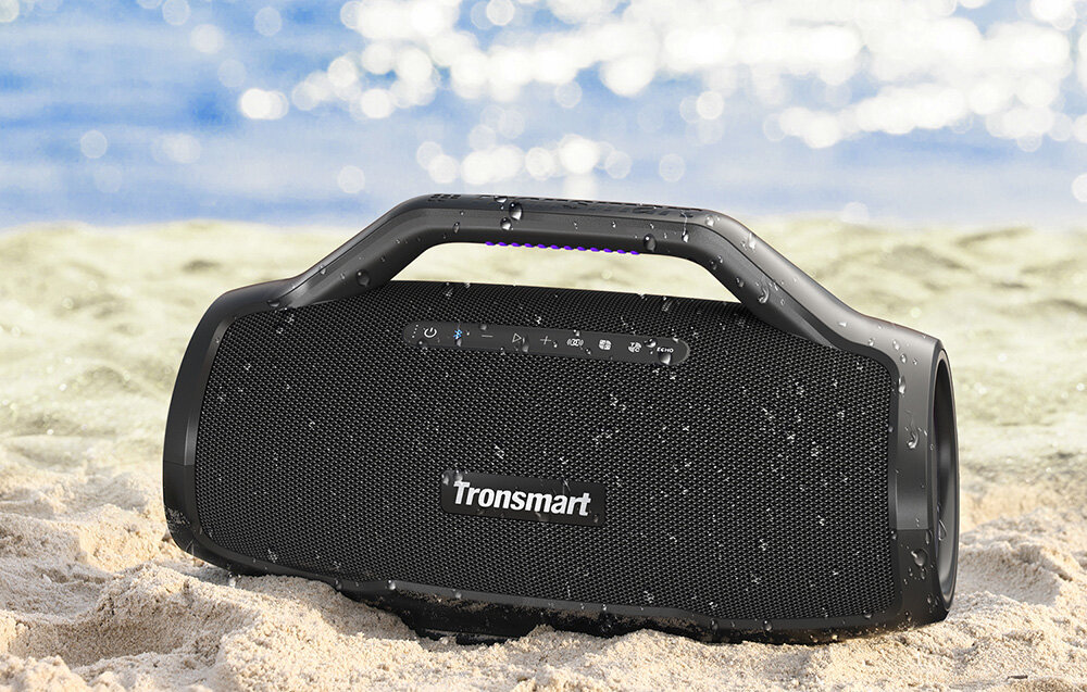 Czarny głośnik Tronsmart na piasku, widoczne krople wody. Tło to rozmyte morze i niebo.