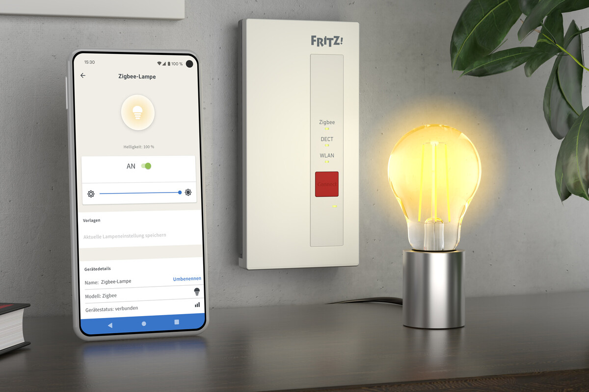Une ampoule allumée sur un support argenté, téléphone avec écran de contrôle de lampe, appareil mural et feuilles vertes.