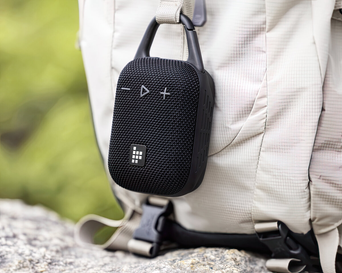 Czarny głośnik wisi na plecaku. Głośnik ma siatkowy przód z przyciskami. Plecak jest beżowy.