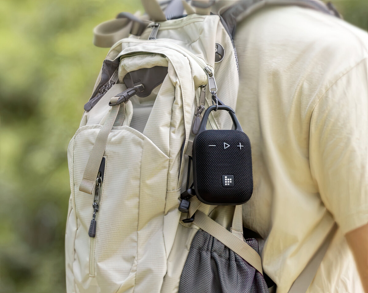 Czarny głośnik przenośny wisi na plecaku. Plecak jest beżowy. Osoba w beżowej koszuli jest w tle.