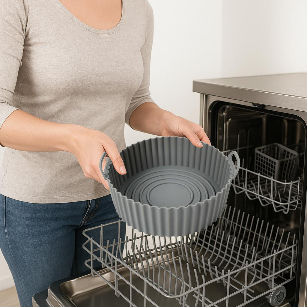 Eine Person hält einen grauen Silikon-Airfryer-Korb in der Nähe einer offenen Spülmaschine. Der Korb ist rund mit geriffelter Kante.