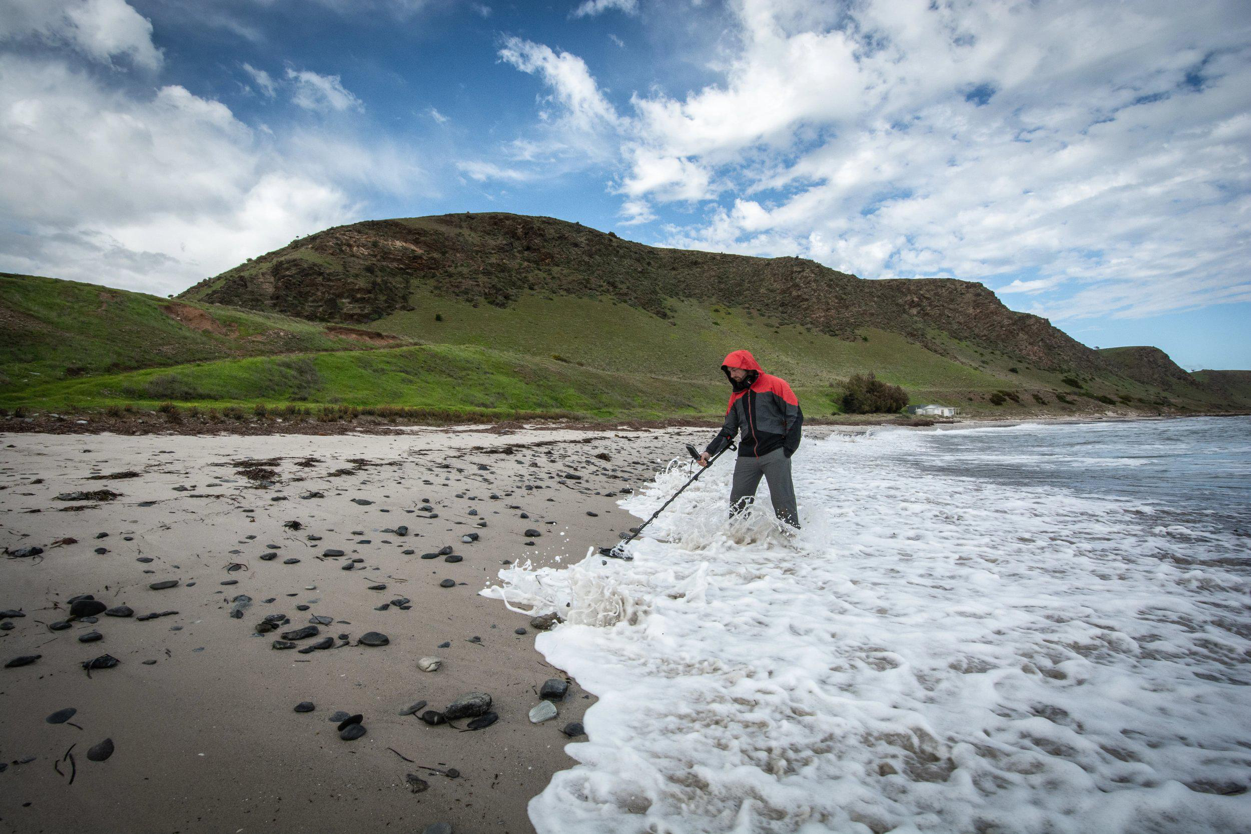 Mann in roter Jacke benutzt Metalldetektor am Strand. Schaumige Wellen und ein grüner Hügel im Hintergrund.