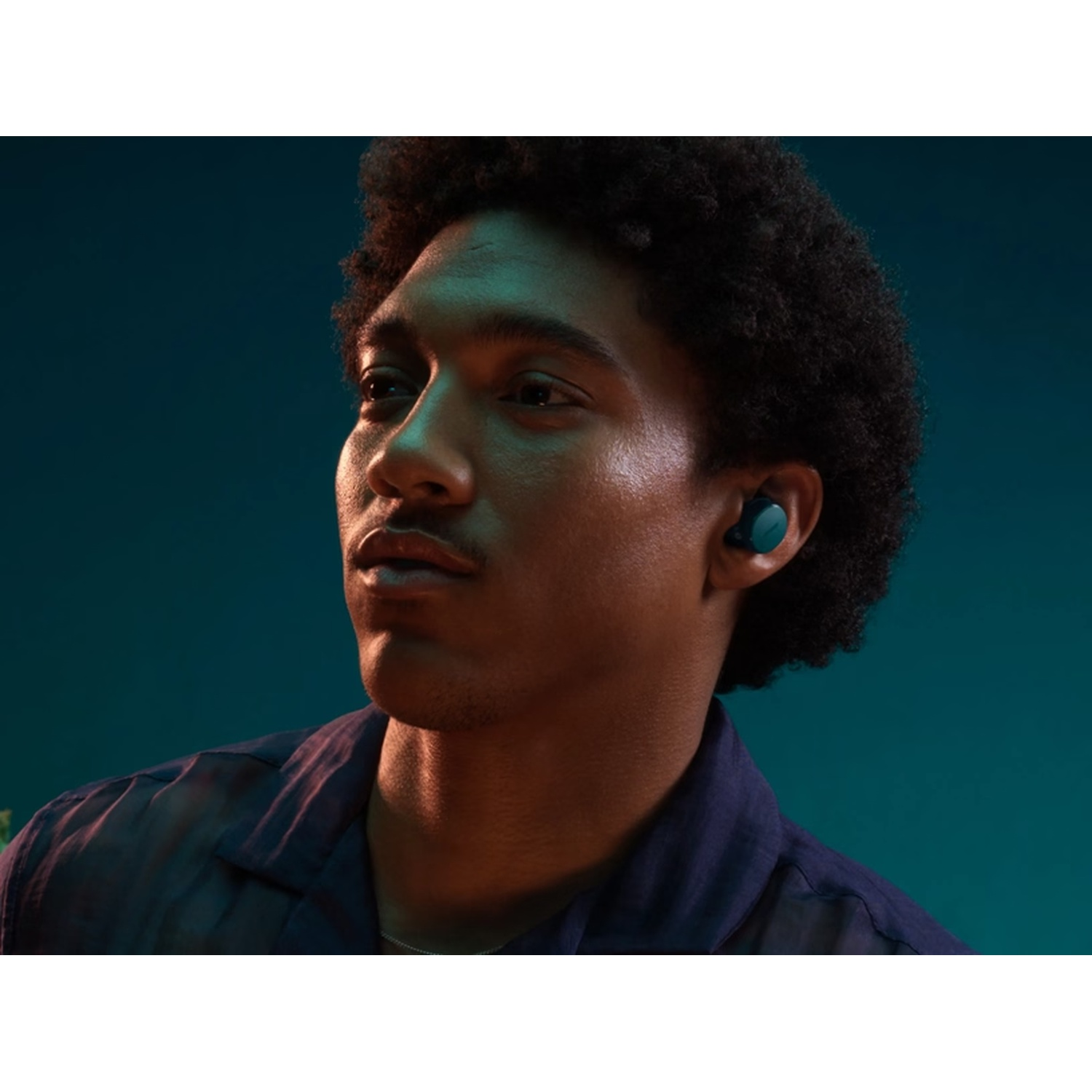 A man with dark skin and curly hair wears an earbud. He is wearing a blue shirt, and the background is a teal color.