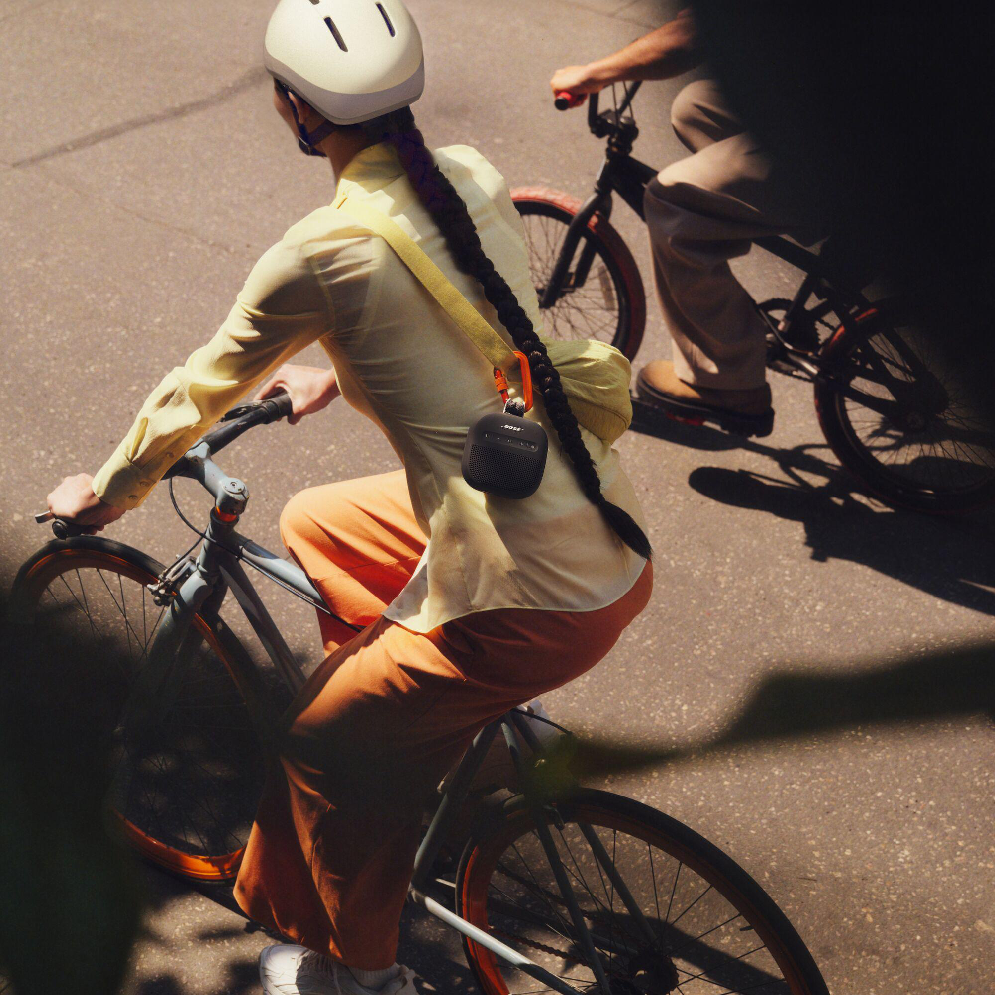 Eine Person fährt an einem sonnigen Tag Fahrrad, trägt einen Helm und ein gelbes Hemd. Ein schwarzer Lautsprecher hängt an der Schulter.
