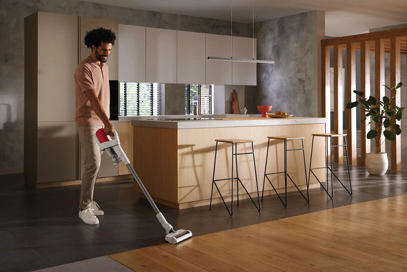 Ein Mann saugt eine moderne Küche. Der Mann trägt ein pfirsichfarbenes Hemd und khakifarbene Hosen. Die Küche hat Holz- und Grautöne.