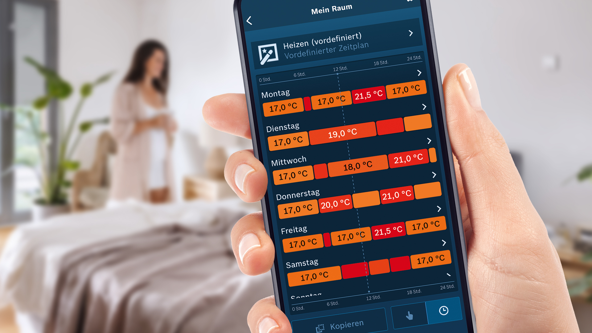 Eine Person hält ein Telefon mit einer Heizungsplan-App. Im Hintergrund steht eine verschwommene Frau in einem Raum.