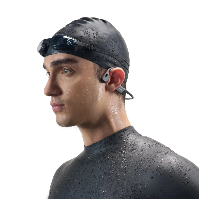 Un hombre con gorro de baño, gafas y auriculares. Mira hacia un lado con expresión seria. El fondo es blanco.