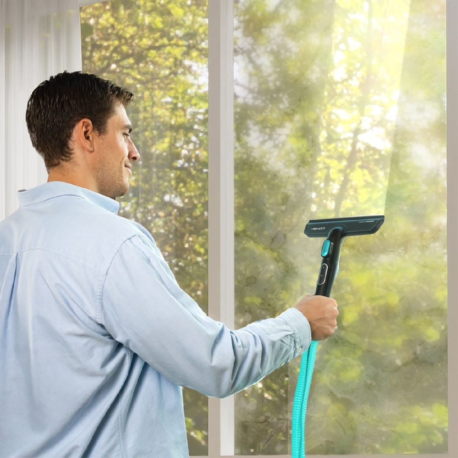 Hombre limpiando una ventana con una escobilla de goma. Viste camisa celeste. El fondo muestra árboles fuera.