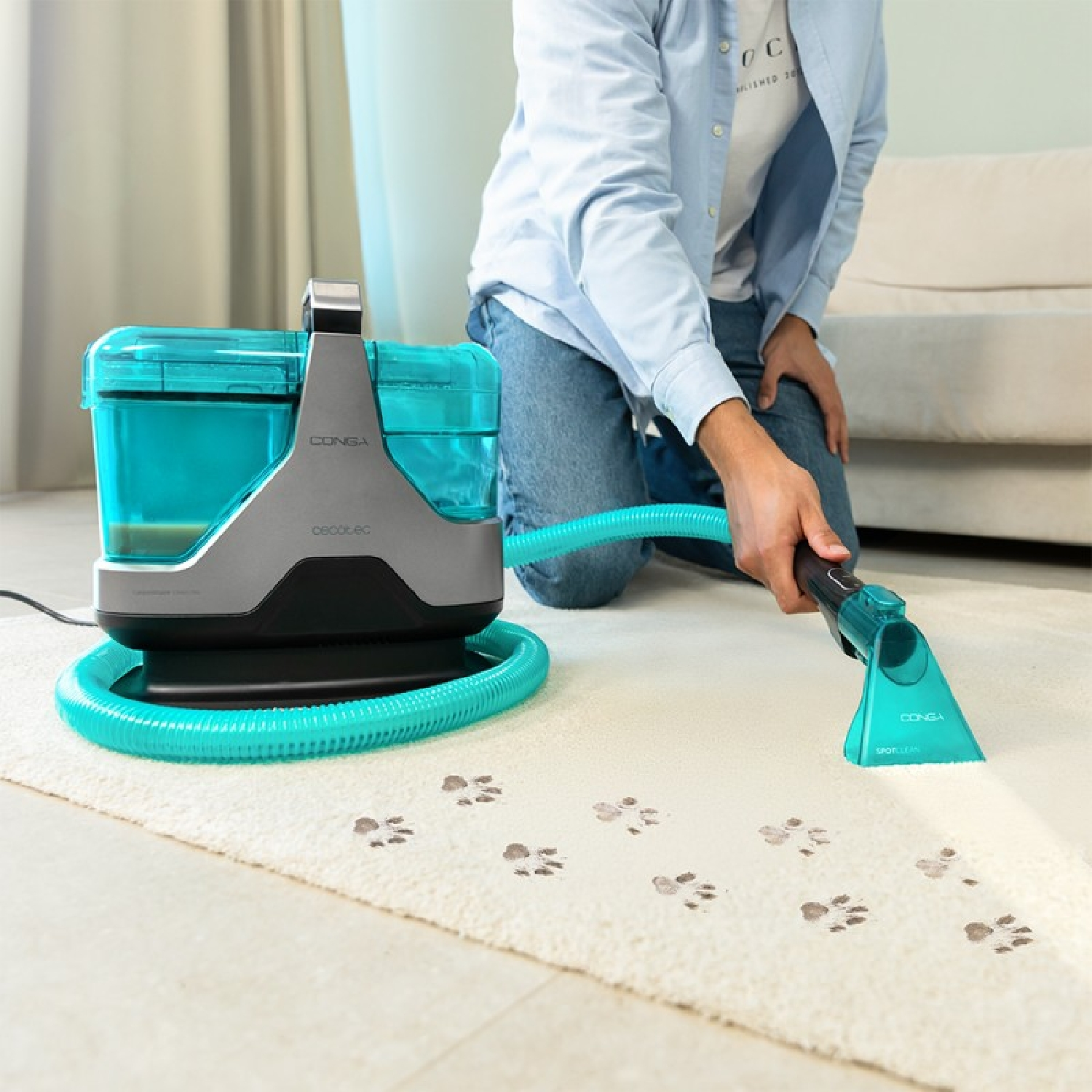 Persona limpiando una alfombra con un limpiador verde azulado, quitando huellas de patas. El limpiador tiene un tanque y una manguera verde azulado.