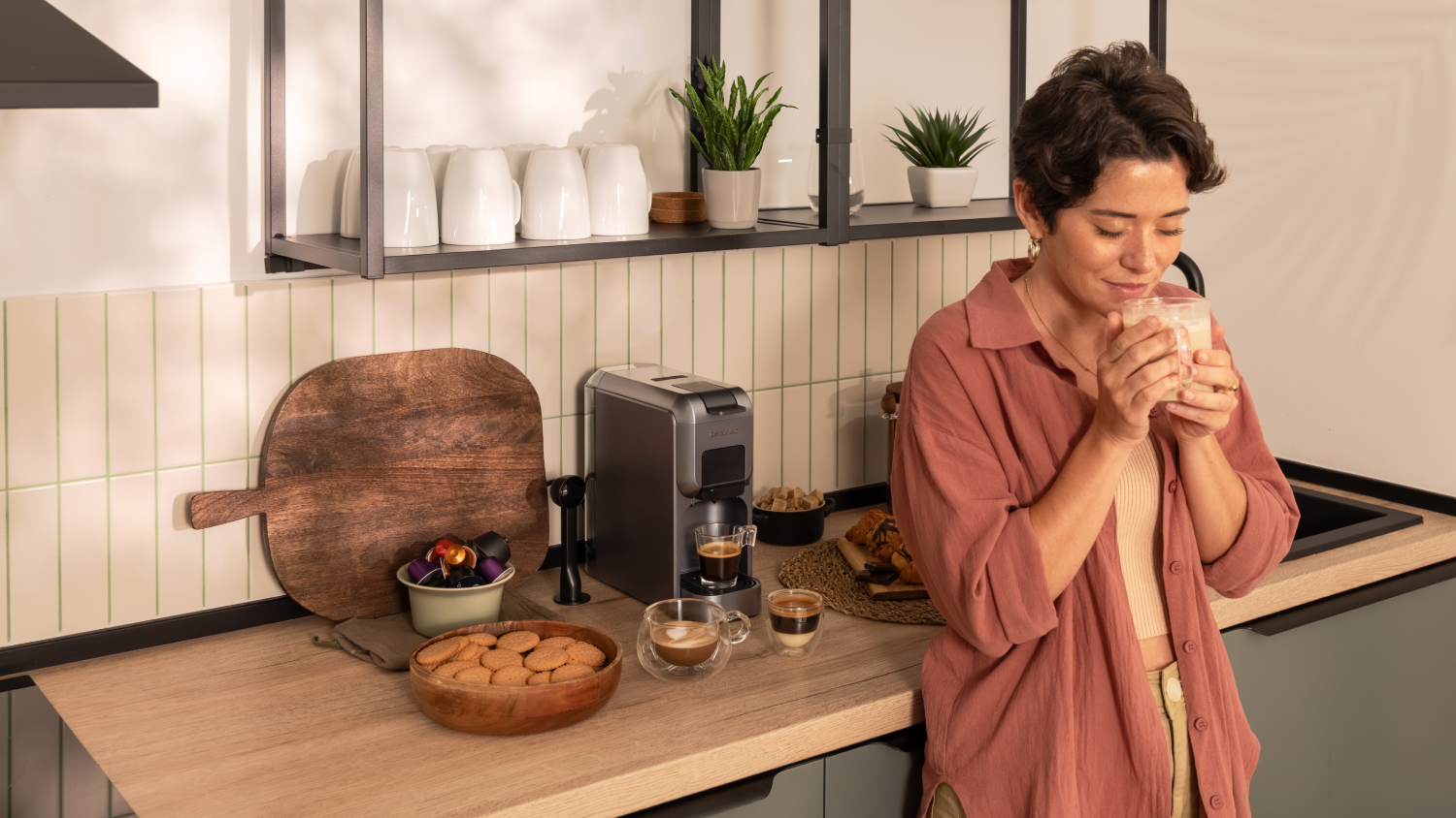 Mujer en la cocina huele café en una taza de vidrio. Cafetera, galletas, plantas y tabla de madera en la encimera.