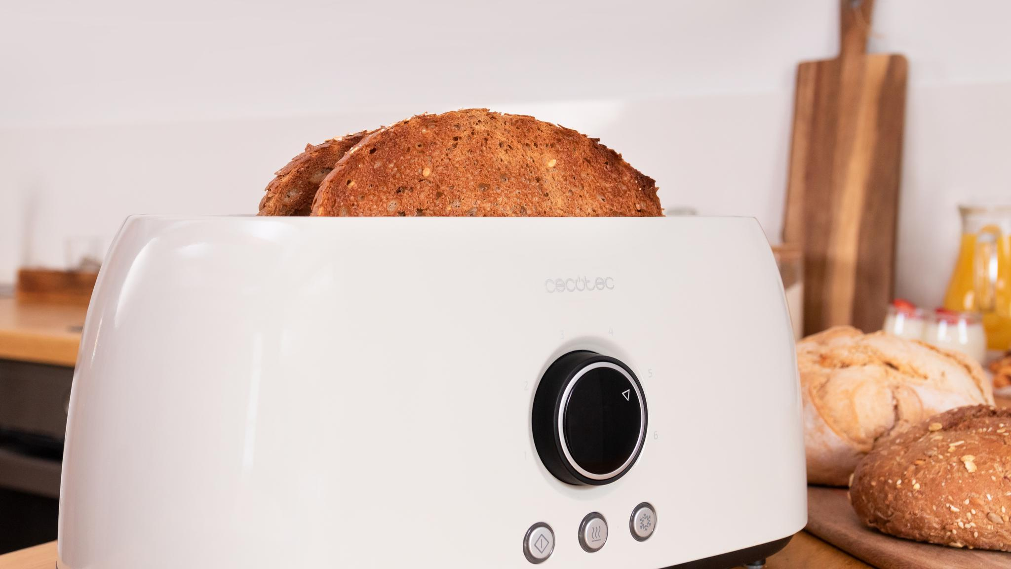 Una tostadora blanca con dos rebanadas de pan tostado. El fondo incluye pan, una tabla de cortar y un recipiente de jugo.