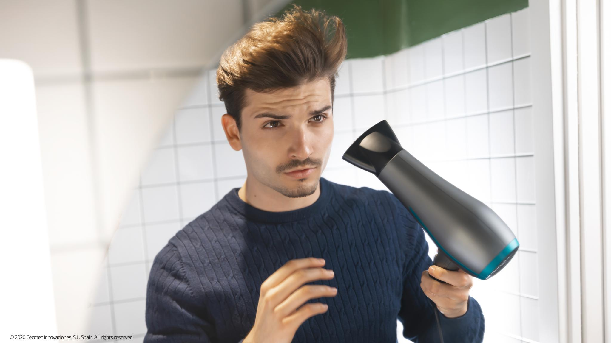 Un hombre con cabello castaño y bigote usa un secador de pelo gris y turquesa. Viste un suéter azul en un baño.