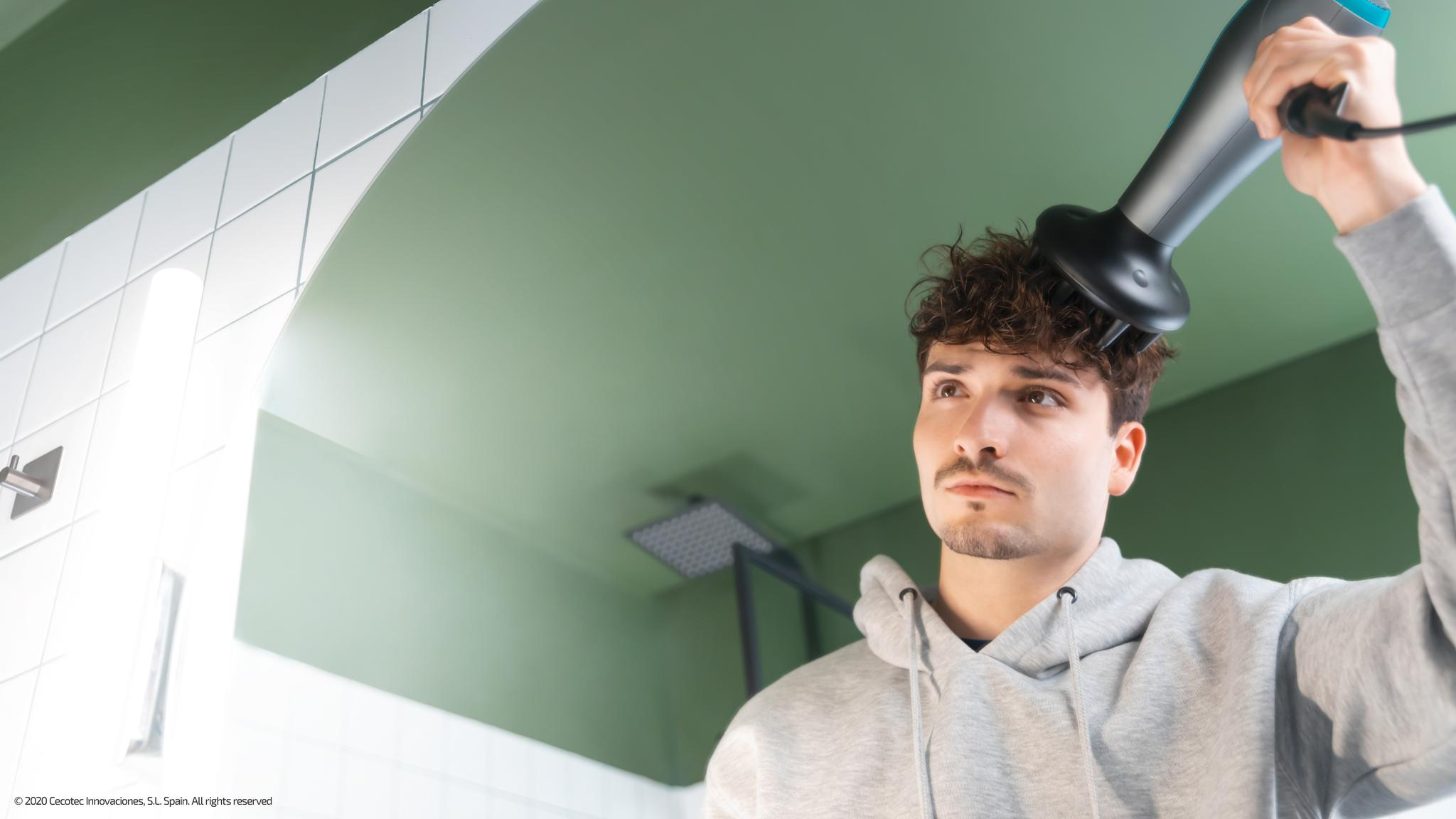 Un hombre se seca el pelo rizado con un secador en un baño. Se ven una pared verde y una pared alicatada blanca. Lleva una sudadera gris.