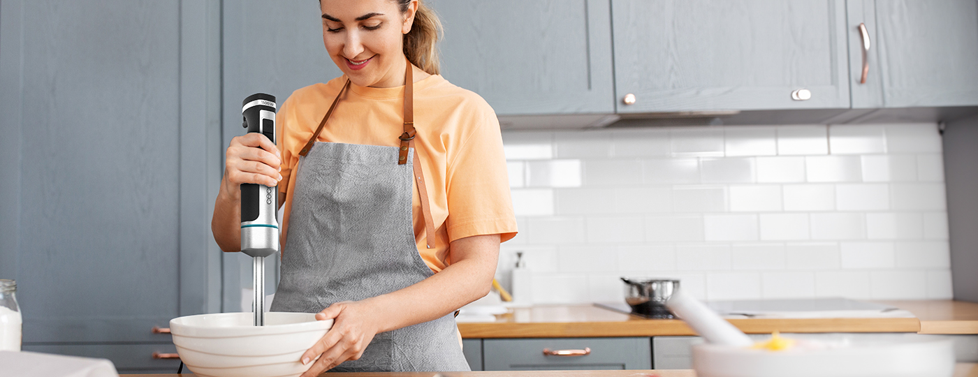 Una mujer con delantal usa una batidora de mano en una cocina. Sonríe. Hay un bol delante de ella.