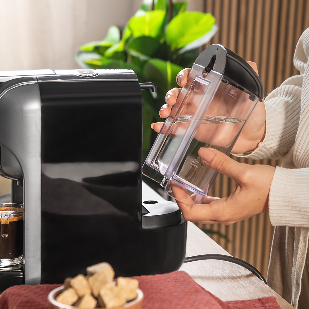 Una persona llena una cafetera negra y plateada con agua de un recipiente transparente. Un café expreso está en un vaso.