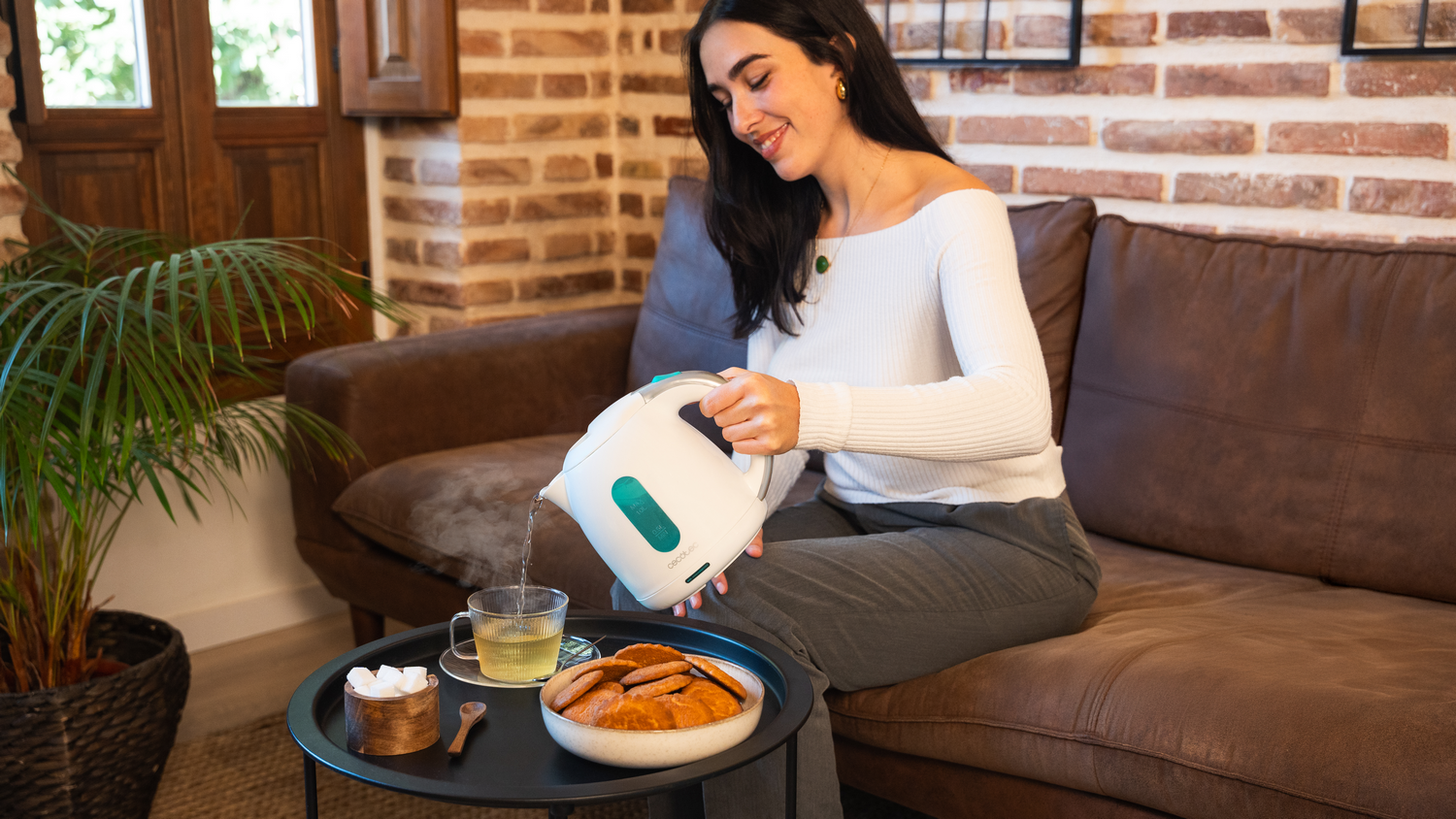 Una mujer vierte agua caliente en una taza de té. Galletas y terrones de azúcar en la mesa. Ella está sentada en un sofá marrón.