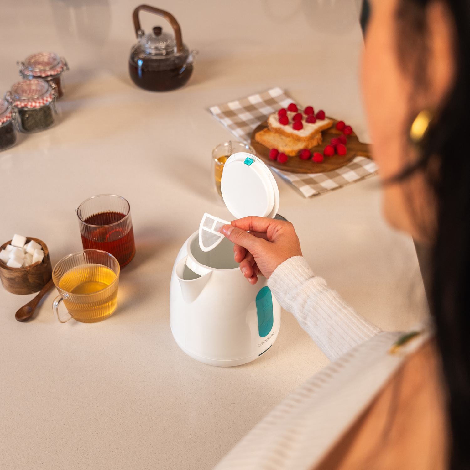 Una persona inserta un filtro en una tetera blanca. Té, tostadas con frambuesas y otros artículos de cocina están en la encimera.
