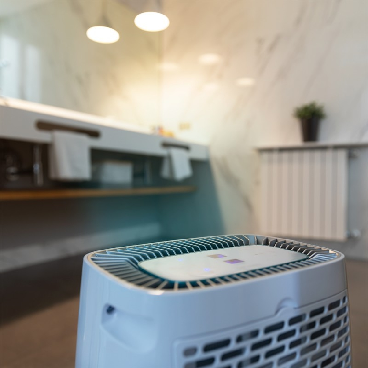 Un purificador de aire blanco en un baño. En el fondo, se ven un lavabo, toallas y una planta. El baño tiene un ambiente luminoso.