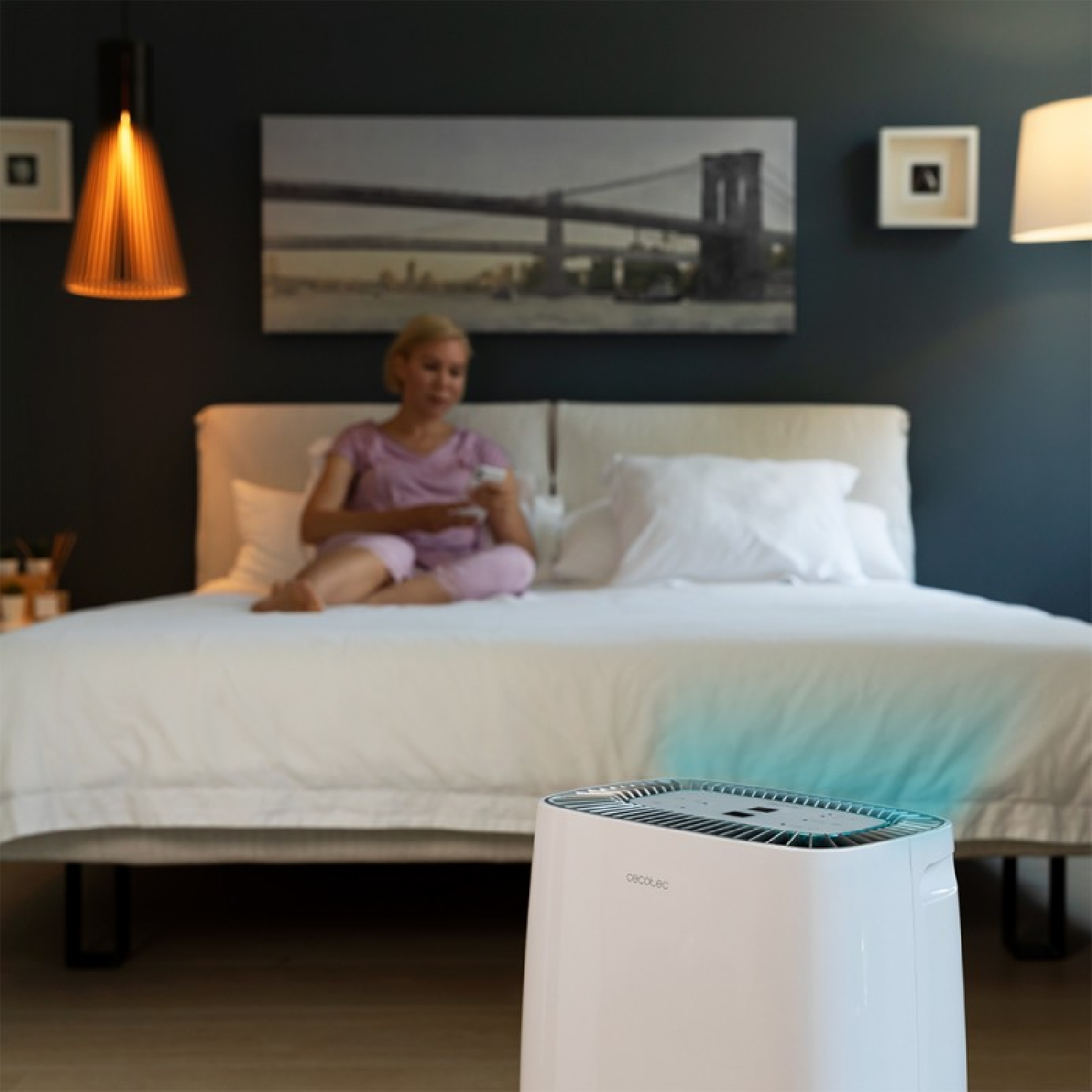 Una mujer sentada en una cama usa un mando a distancia. Un purificador de aire blanco está en primer plano. Se ven un cuadro y lámparas.