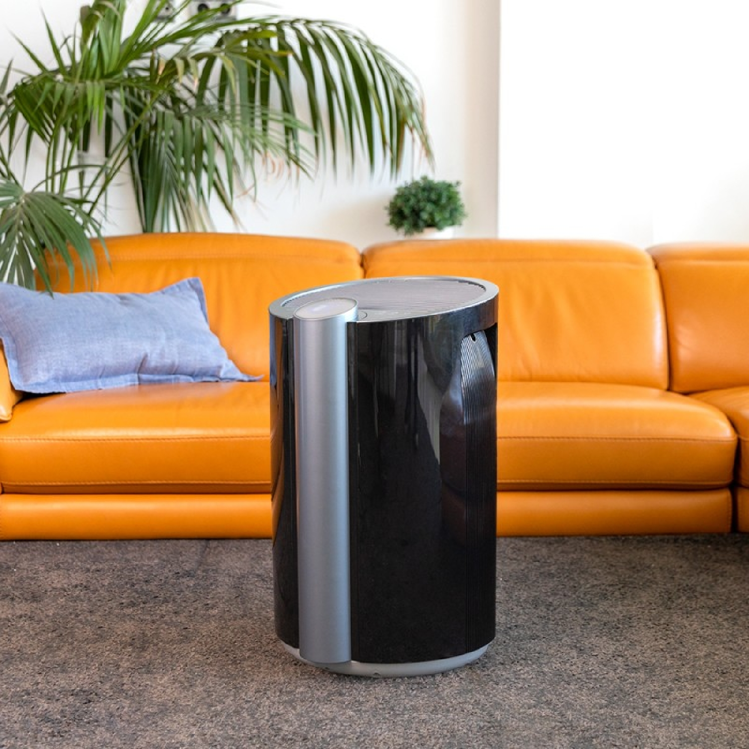 Un purificador de aire negro frente a un sofá naranja. Una almohada azul en el sofá. Una planta a la izquierda.