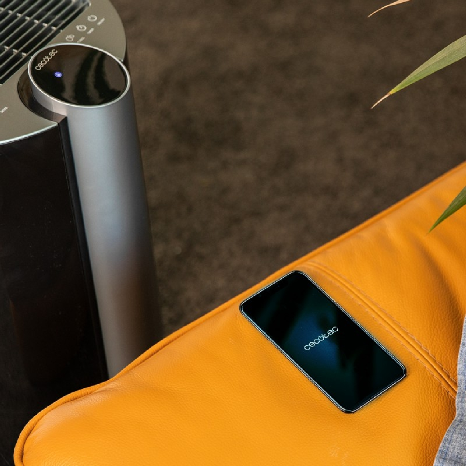 Un purificador de aire, teléfono en cojín naranja. El teléfono muestra 'cecotec'. El fondo es marrón, con hojas verdes.