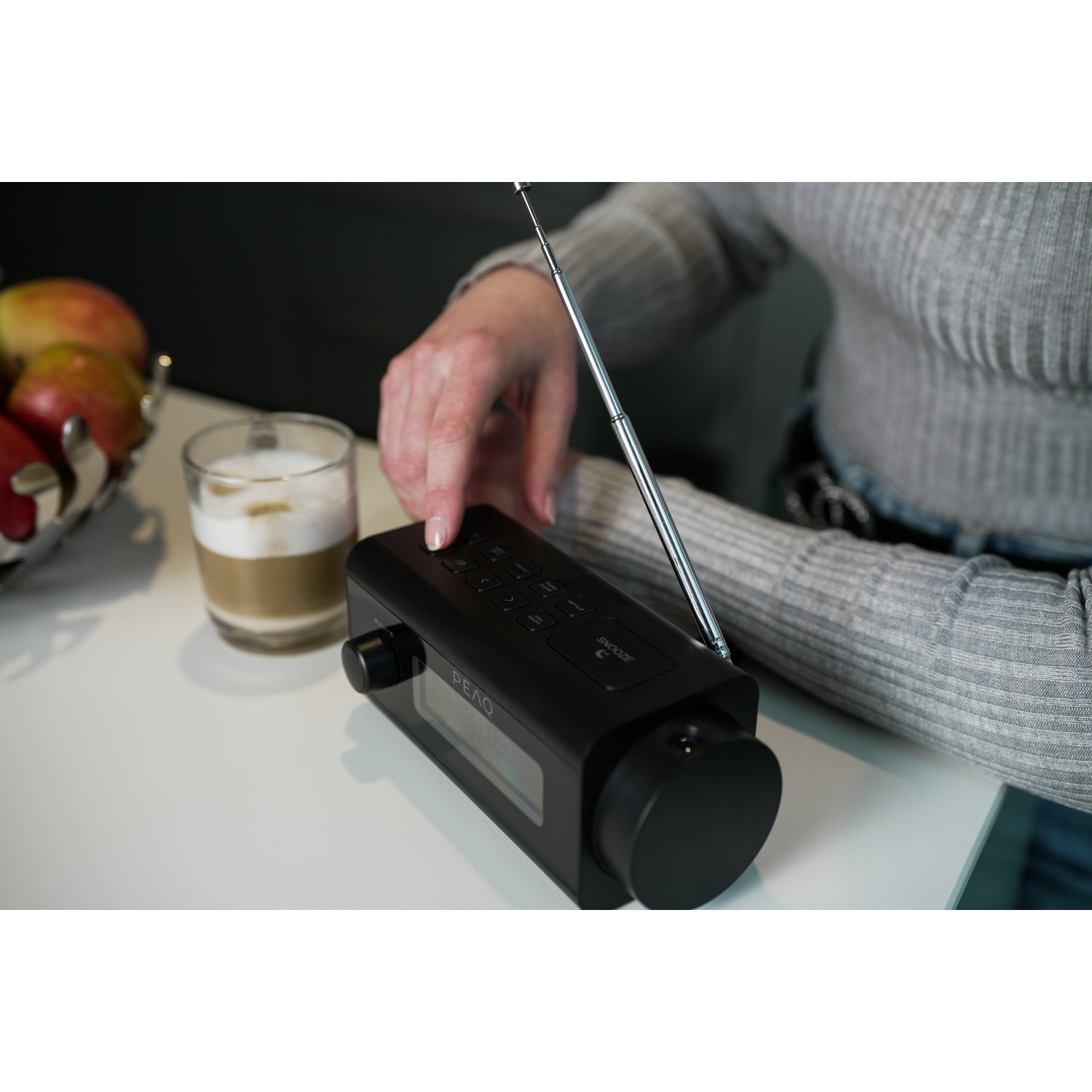A person's hand presses a button on a black radio. A glass of coffee and a fruit bowl are also on the white table.