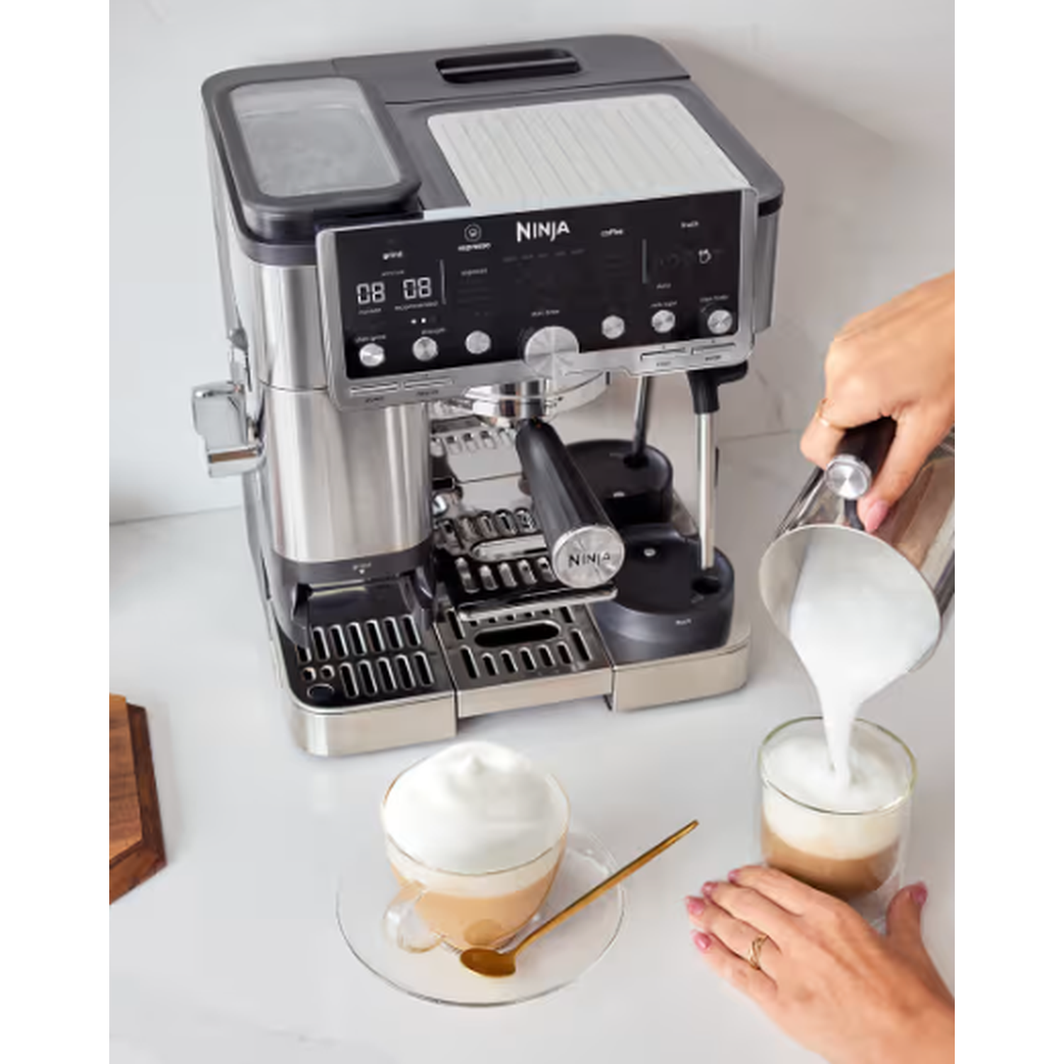 Een Ninja koffiezetapparaat met koffie die in een glas wordt gegoten. Een kopje koffie met schuim staat op tafel. Een hand giet melk.
