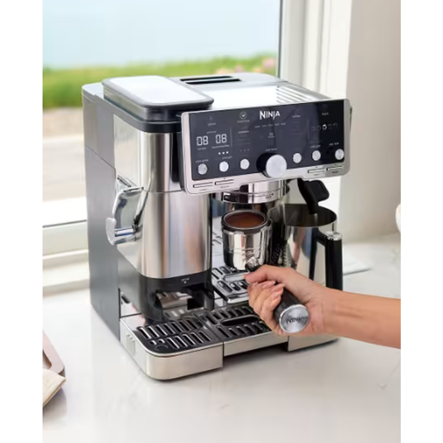Een Ninja espressomachine staat op een aanrecht. Een hand plaatst de filterhouder. De achtergrond toont een raam en de zee.